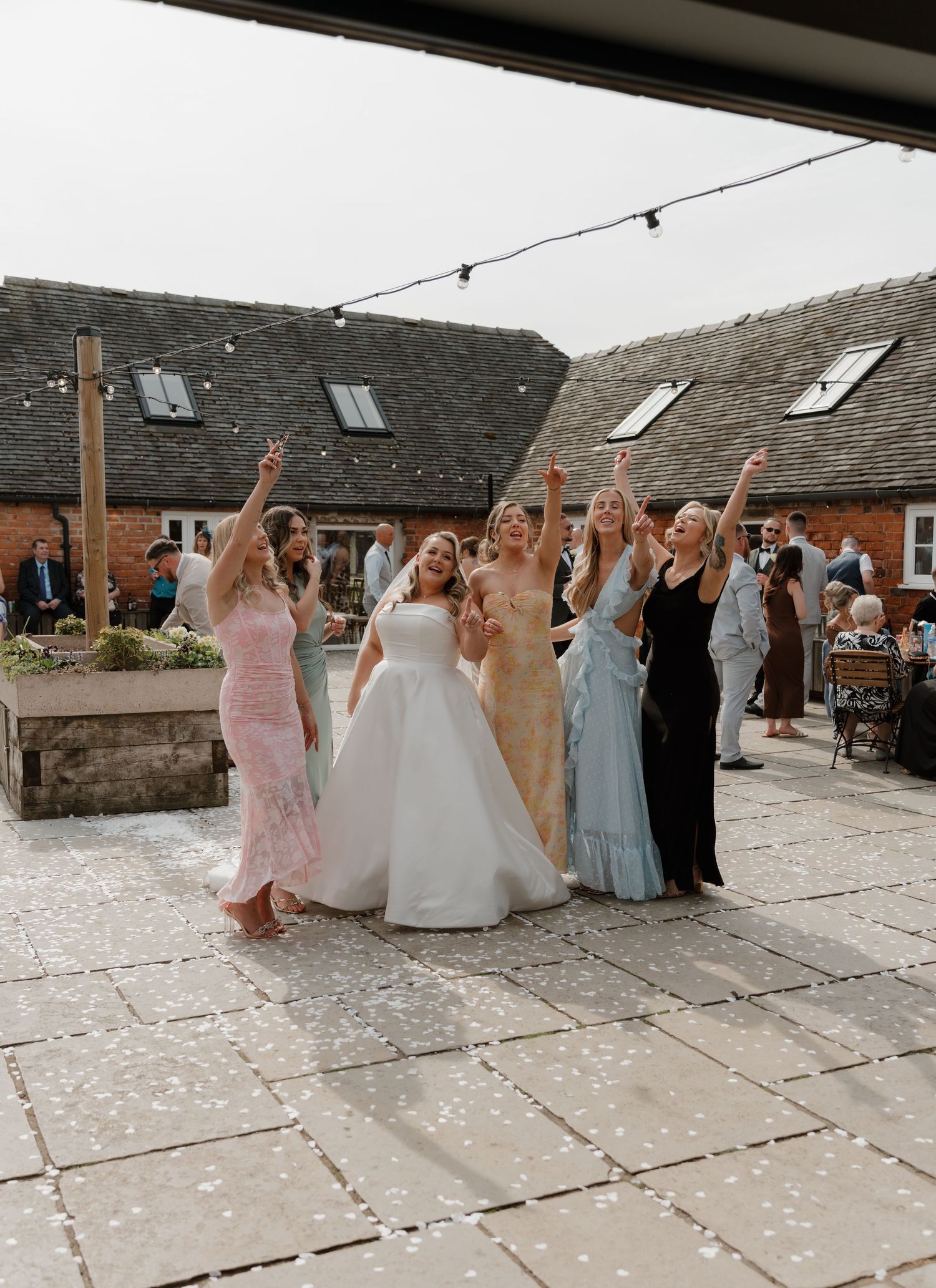 Bride in white gown celebrates with bridesmaids in colorful dresses at outdoor wedding venue with confetti.