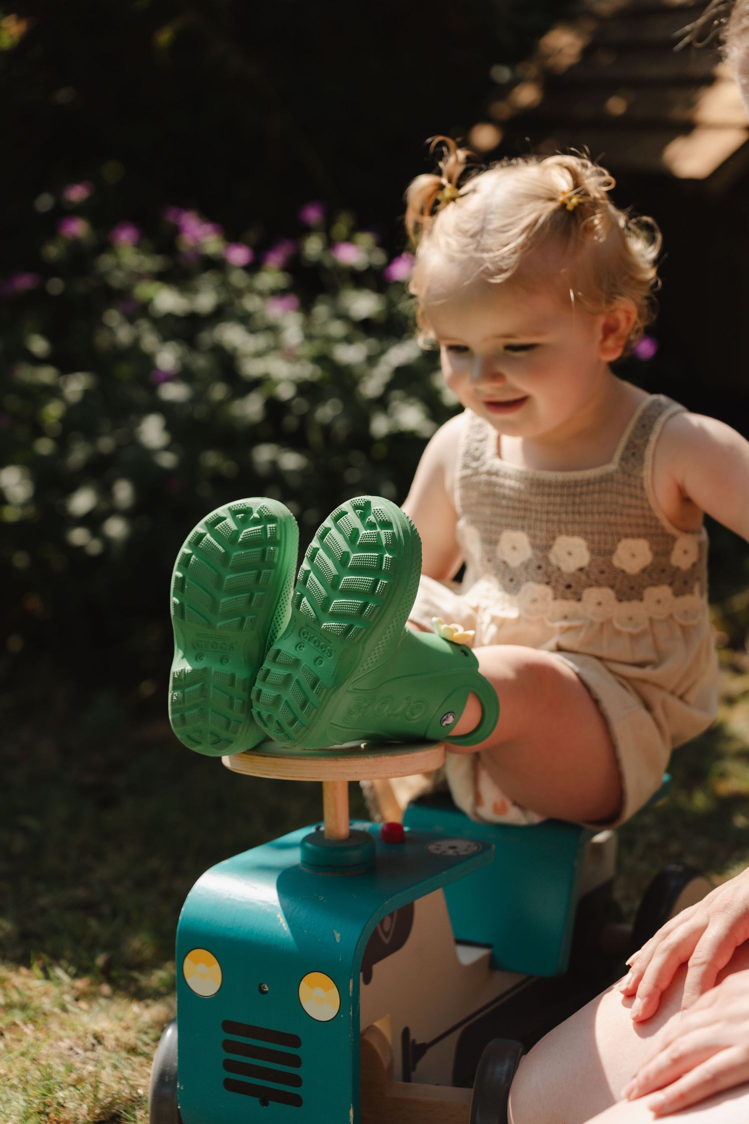 Smiling toddler girl in cream romper sitting on toy tractor wearing green rubber rain boots outdoors.