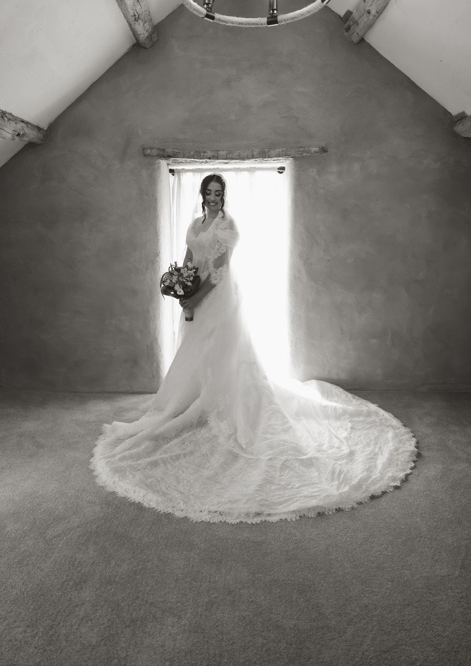Black and white bridal portrait of a bride in a flowing gown with long train, holding bouquet by a bright window.