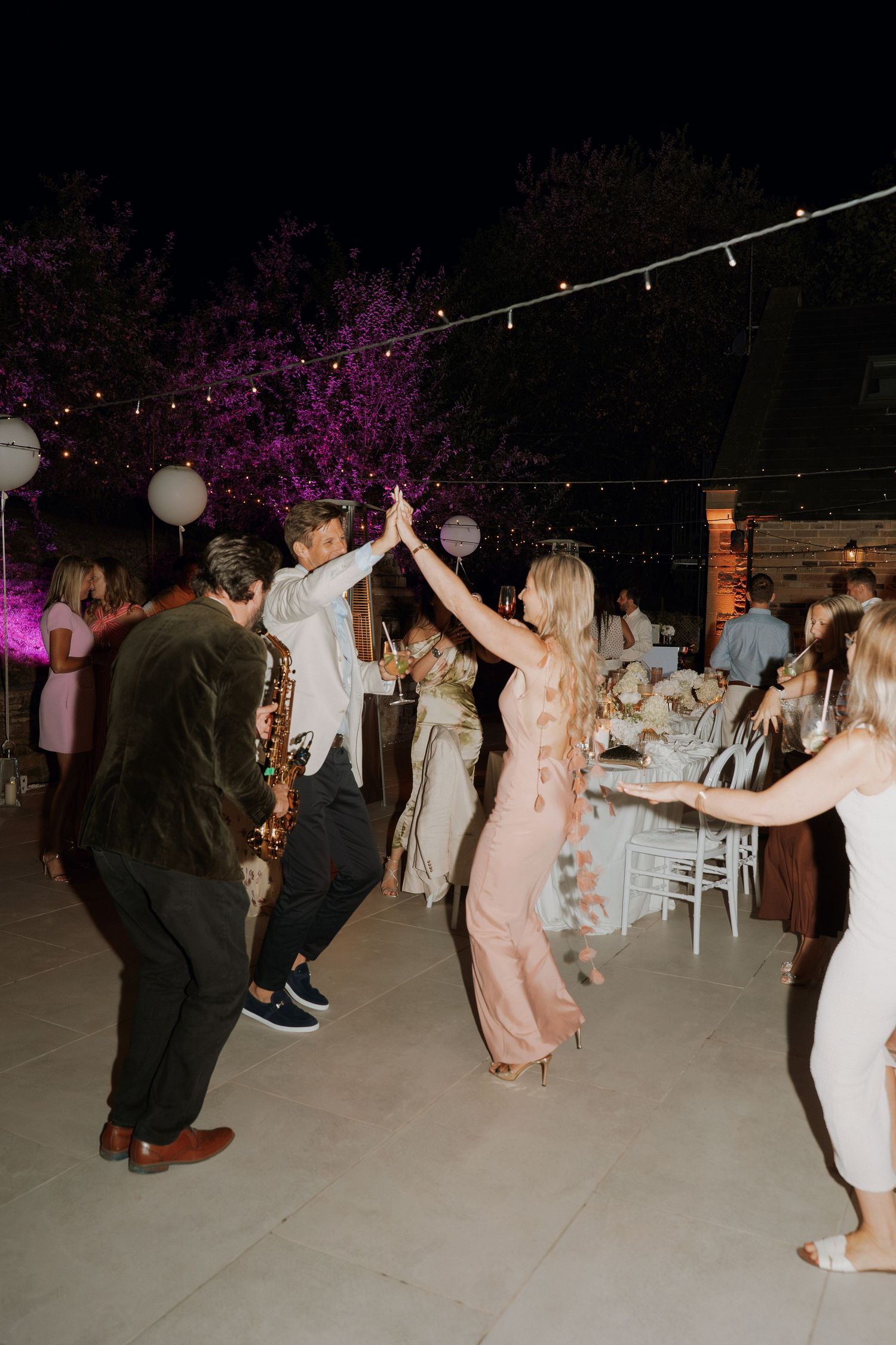 Guests dancing and high-fiving at an outdoor wedding reception at night with string lights and purple-lit trees.