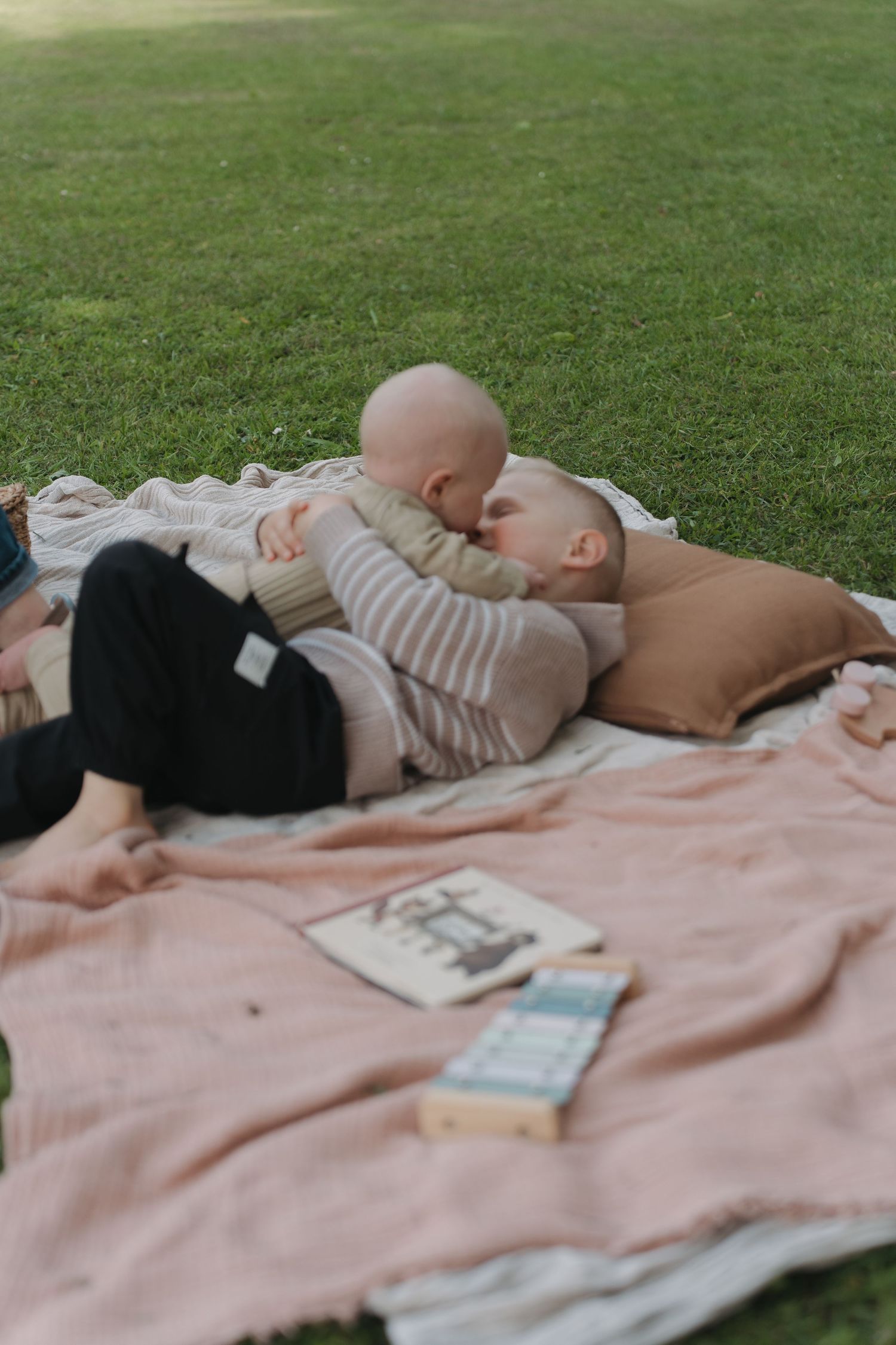 Two people sharing a tender moment on a pink blanket in a park, with books nearby on a sunny day.