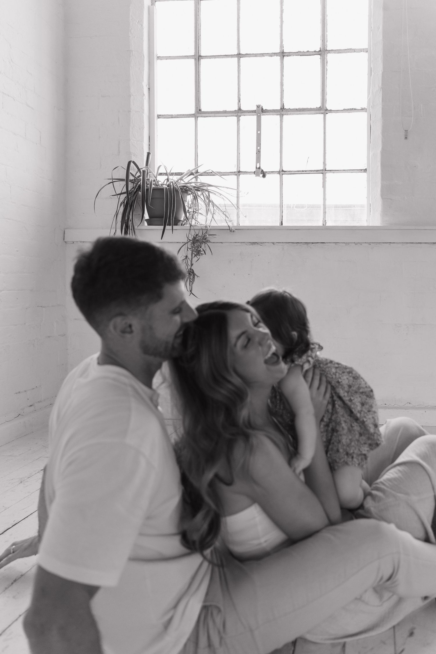 Black and white photo of a couple sitting on the floor with a child in a bright room with large windows.