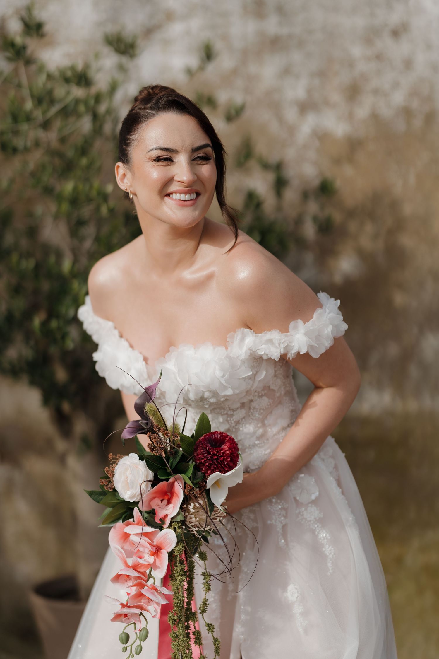 Smiling bride in off-shoulder white gown holding cascading pink floral bouquet outdoors.