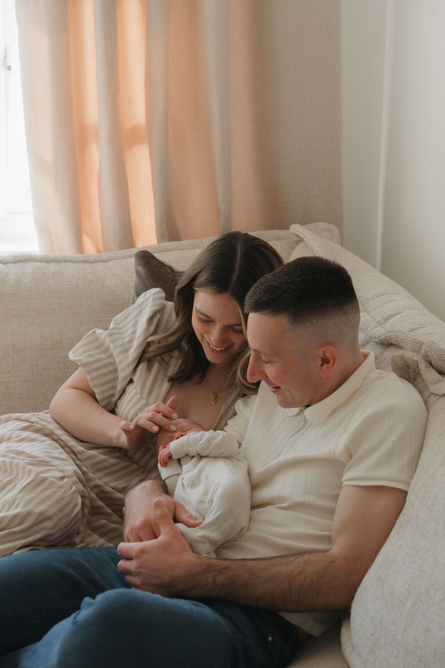 Home Newborn Photography Derby. Happy couple sitting on a cozy sofa, lovingly cradling their newborn baby in a warm, softly lit living room.