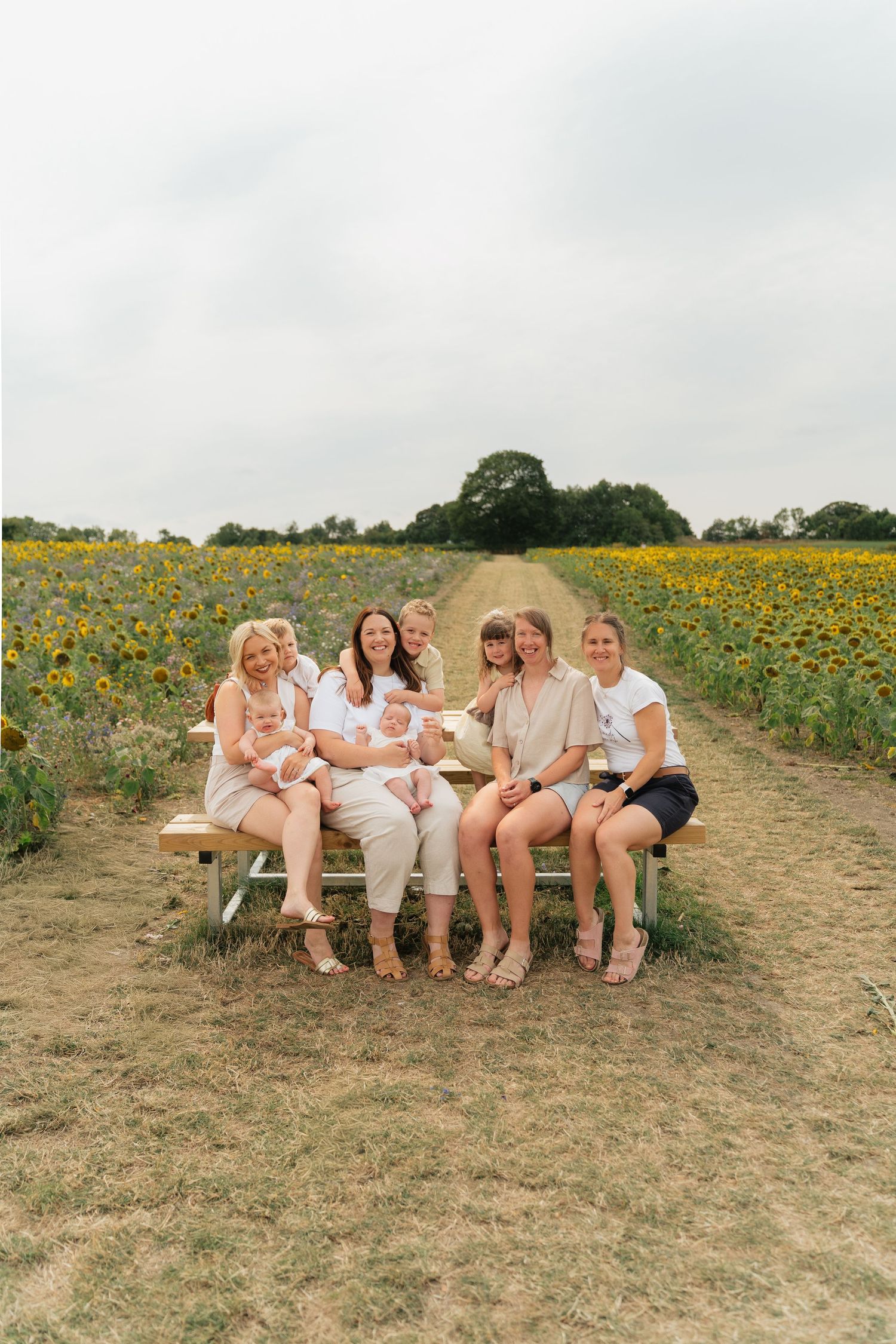 A group in white outfits sits together on a grassy hill overlooking a sunflower field on a cloudy summer day.