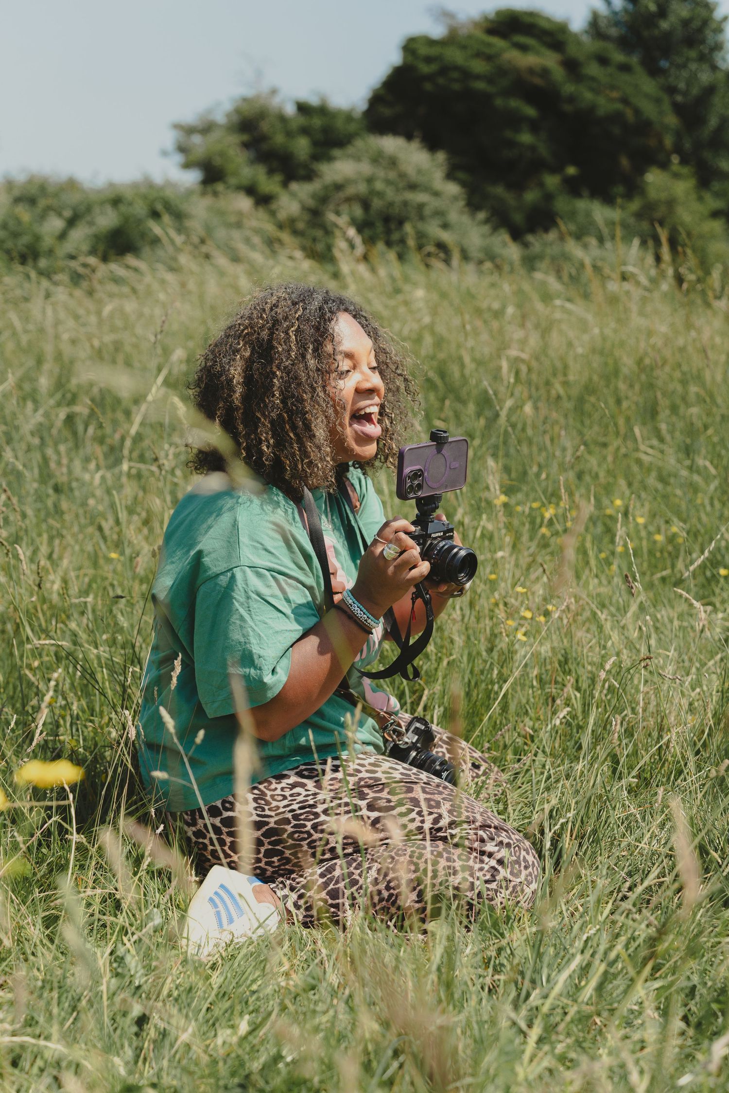 Person in green shirt and leopard print pants laying in tall grass while taking photos.