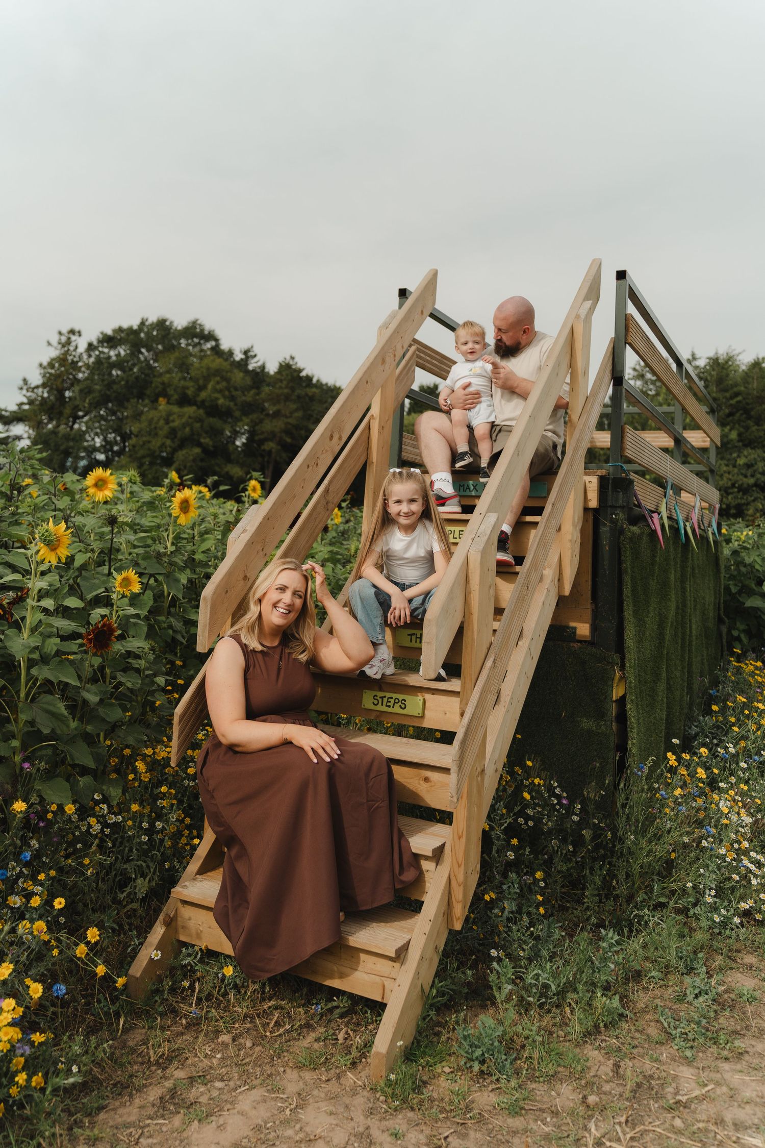 Family of four poses on wooden stairs surrounded by sunflowers, mom in brown dress, dad holding toddler, young girl sitting on steps.