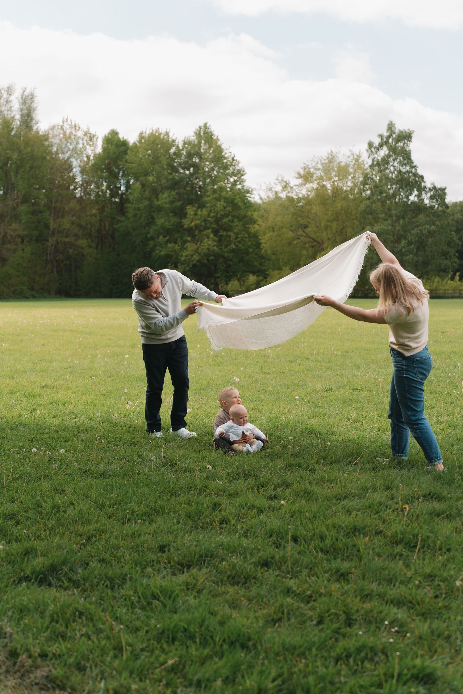 Parents holding a white blanket over their baby sitting on green grass in a park on a cloudy day.