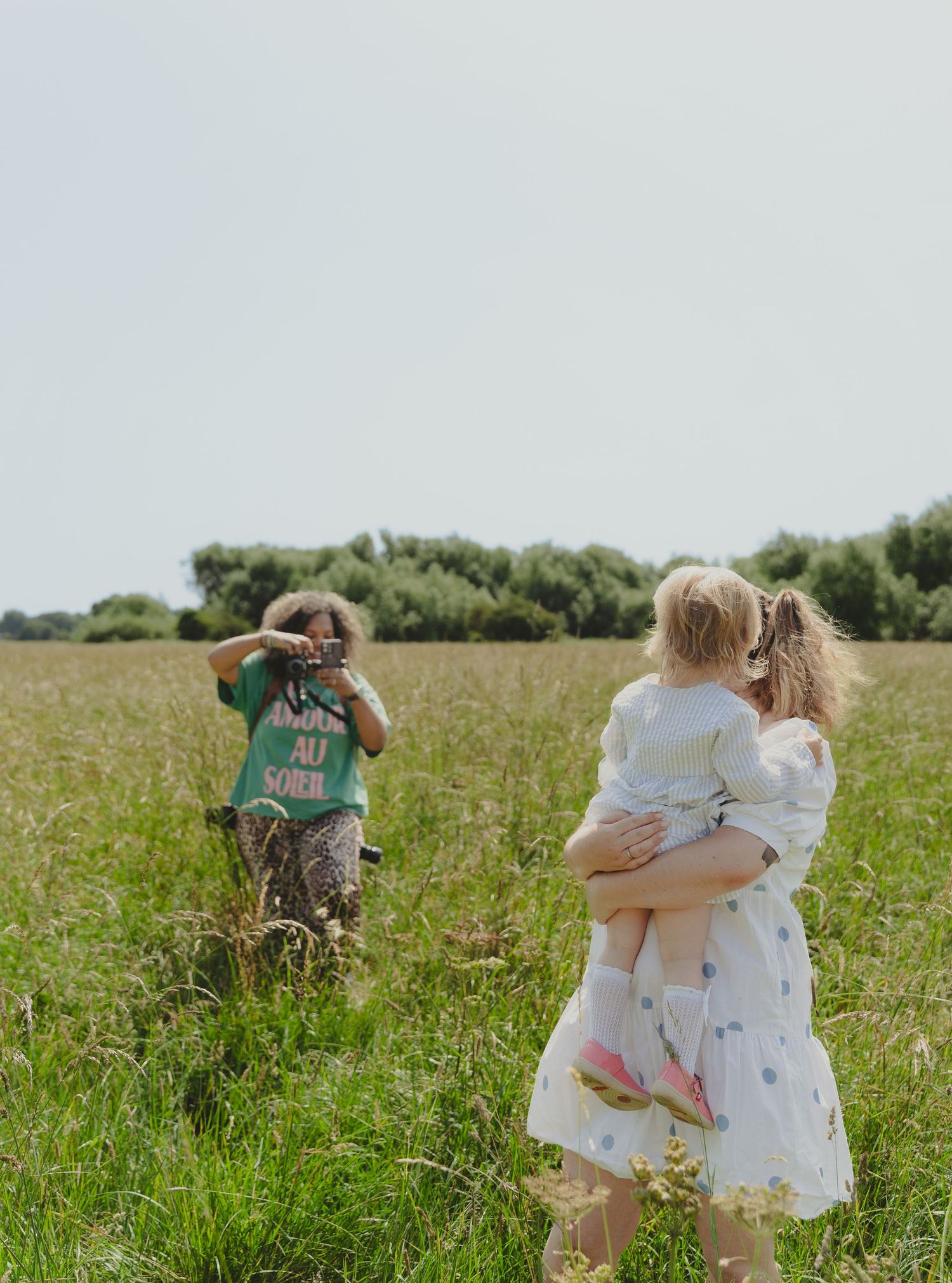 People enjoying a summer day in a grassy meadow with one taking photos while another carries a child.