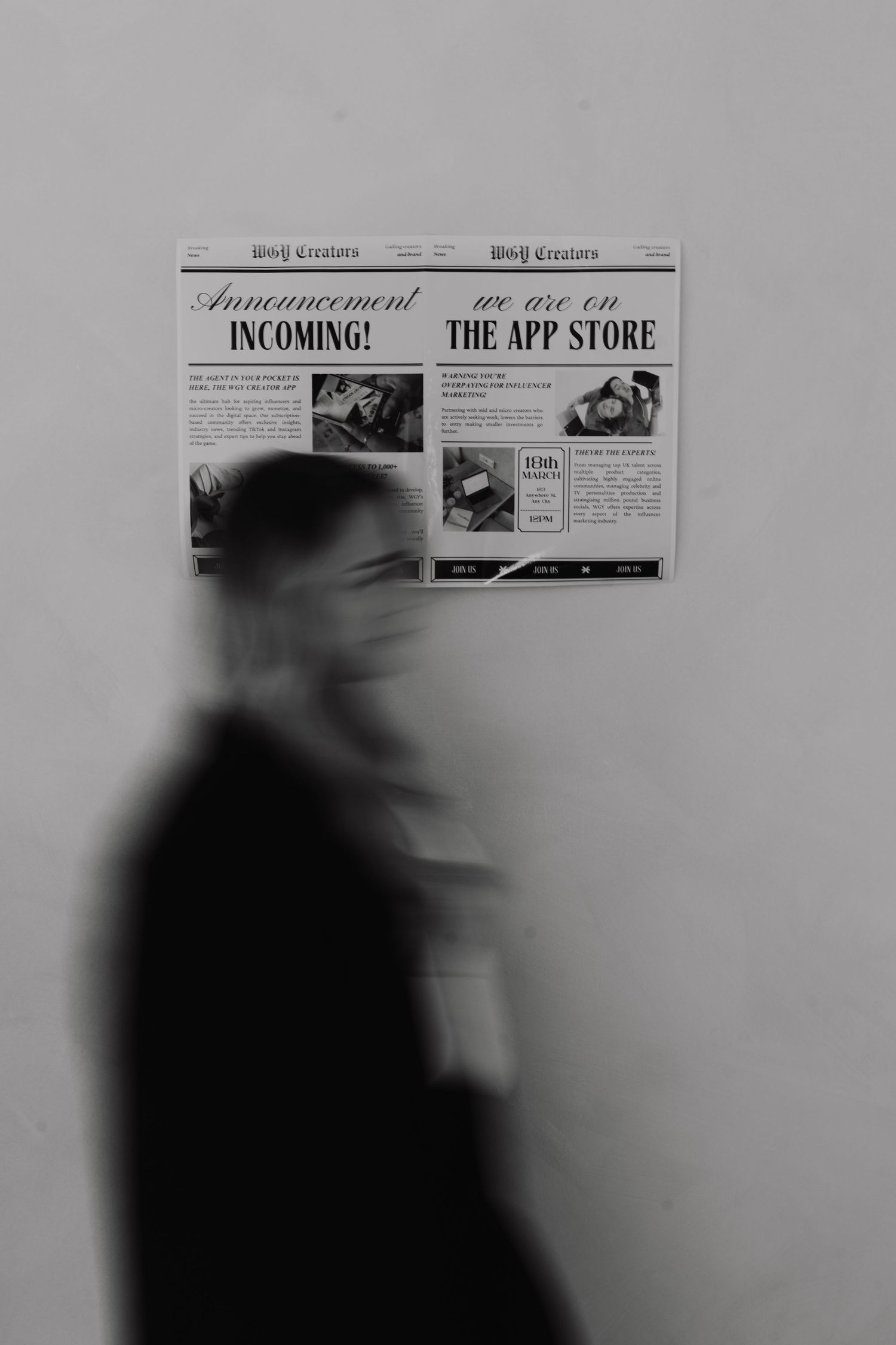 Blurred black and white image shows a silhouetted figure walking past newspaper advertisements on a wall.