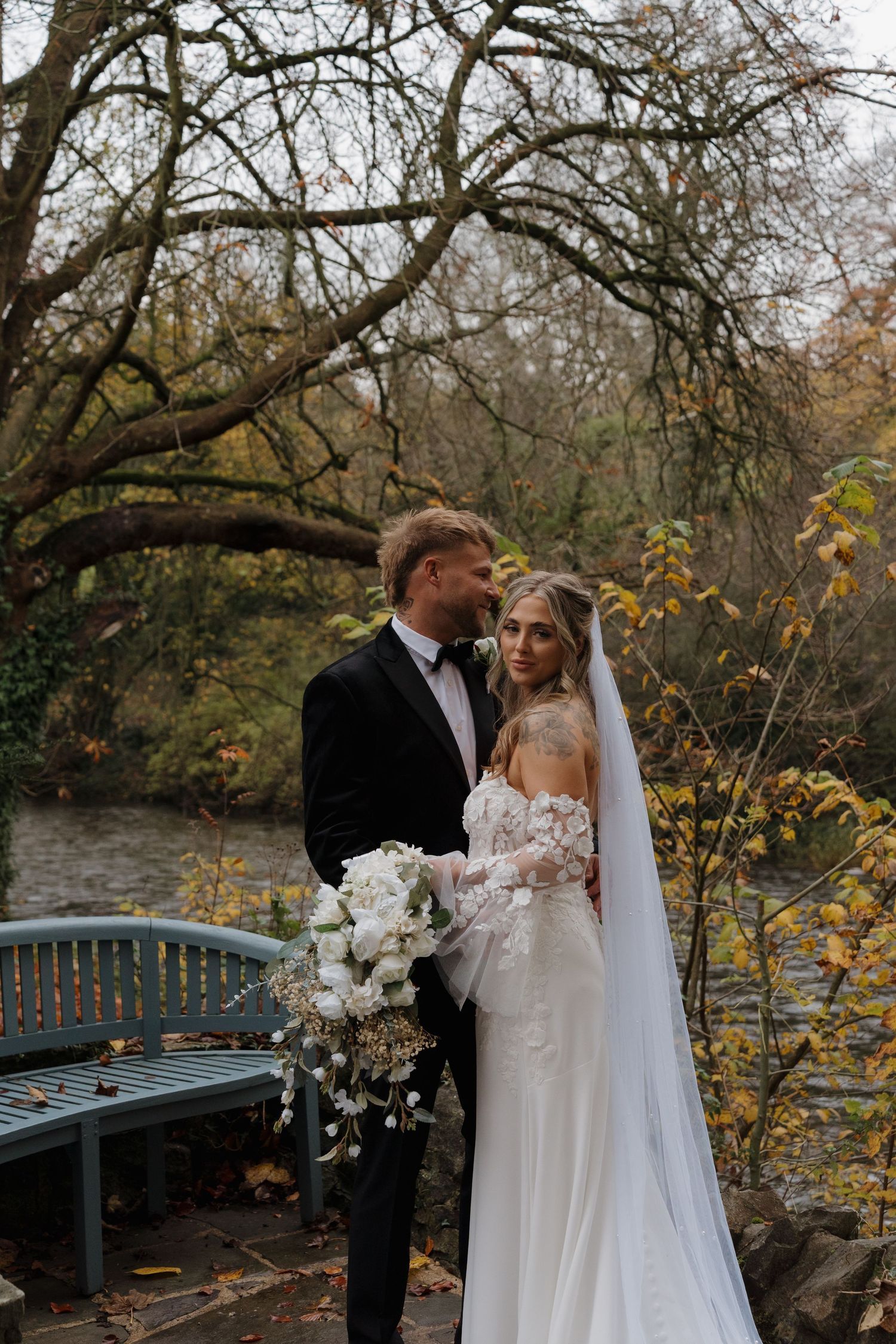 Couple in wedding attire embrace on a rustic bridge surrounded by autumn trees and fallen leaves near a stream.