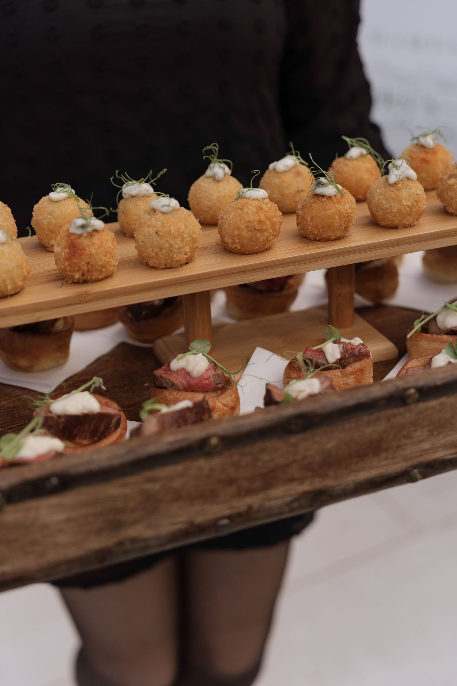 Elegant catering tray with fried arancini balls and bruschetta bites served on a rustic wooden tiered stand.