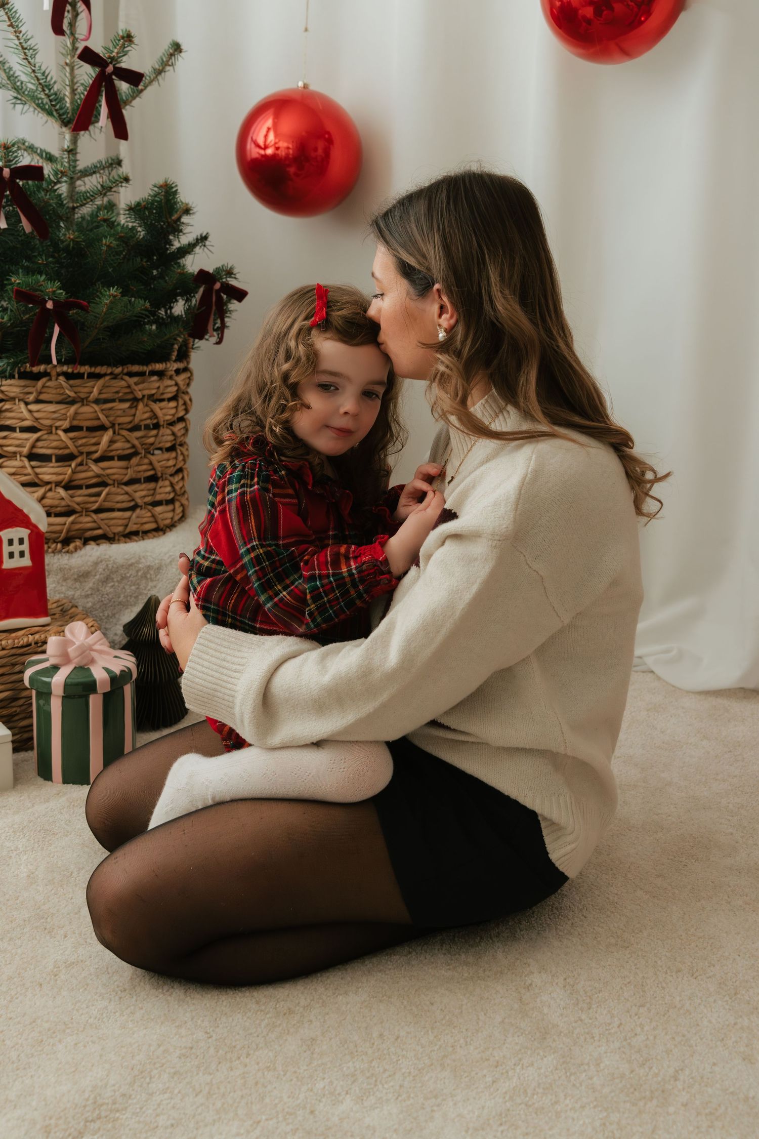 Mother and daughter in Christmas outfits sitting together near decorated tree with red ornaments and gifts.