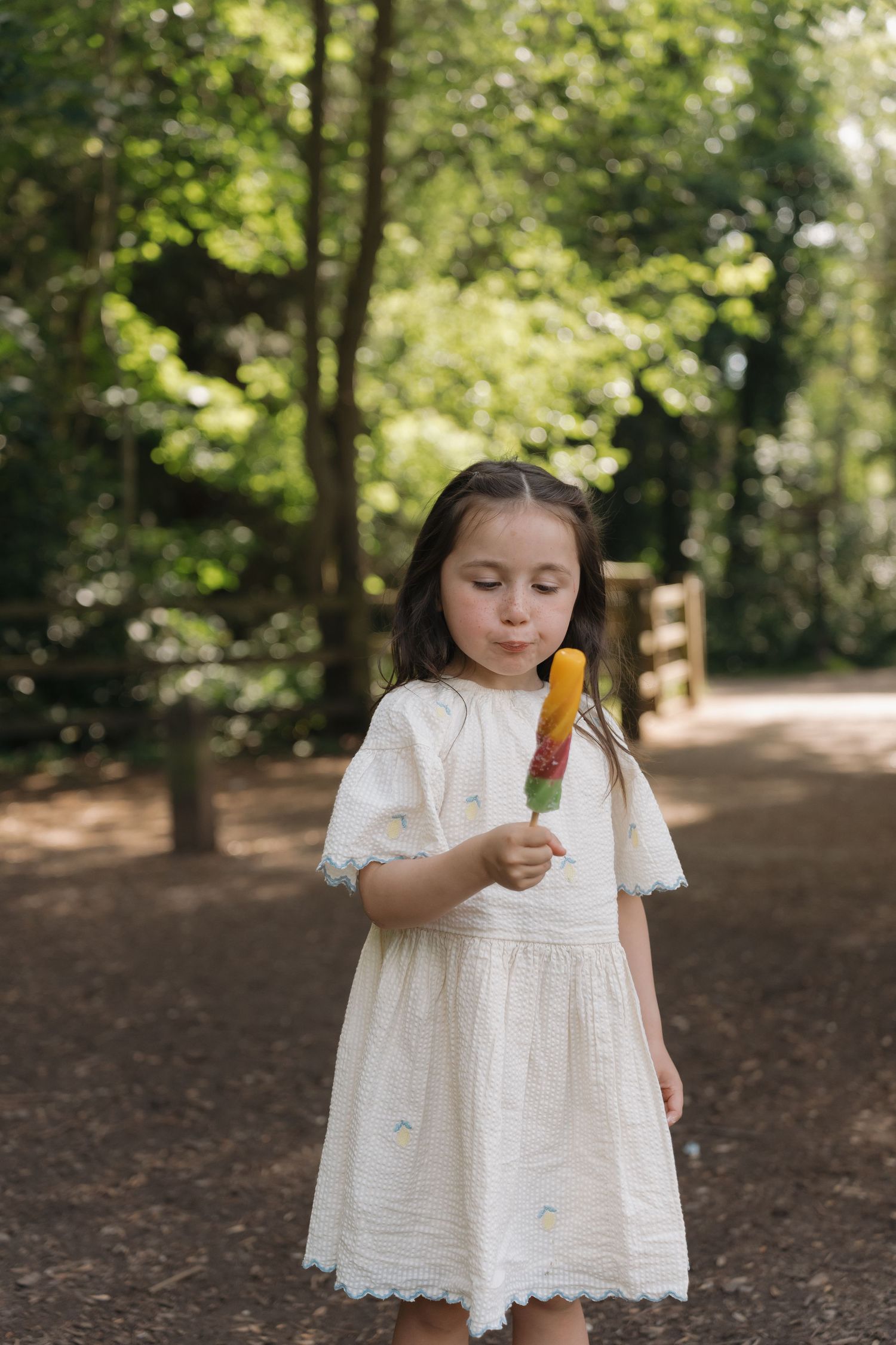 A young girl in a white dress enjoys a colorful popsicle outdoors on a sunny day surrounded by lush green trees.