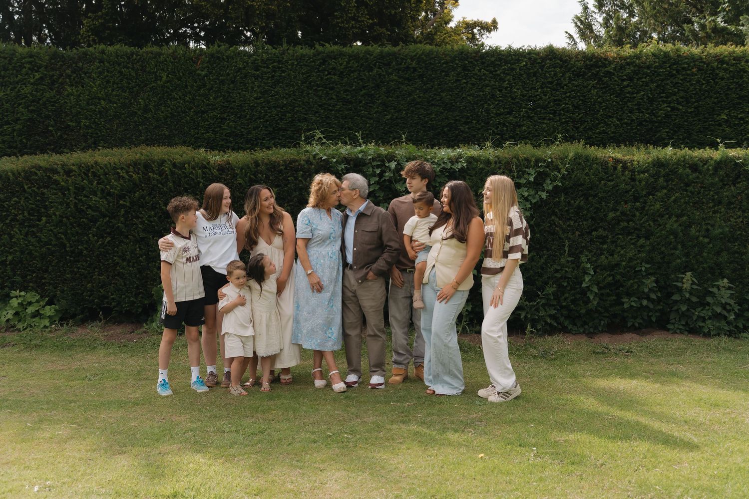 Large multigenerational family group posing together on green lawn in front of tall hedge on sunny day.