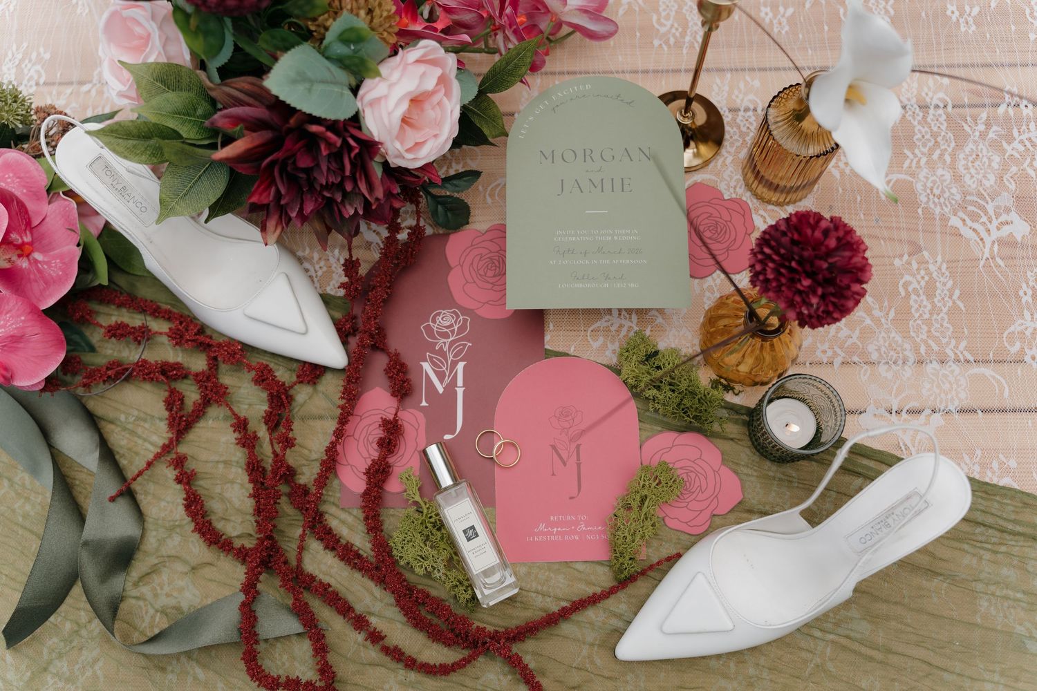 Wedding details flatlay with pink and burgundy florals, white shoes, invitation suite and decorative elements on peach background.