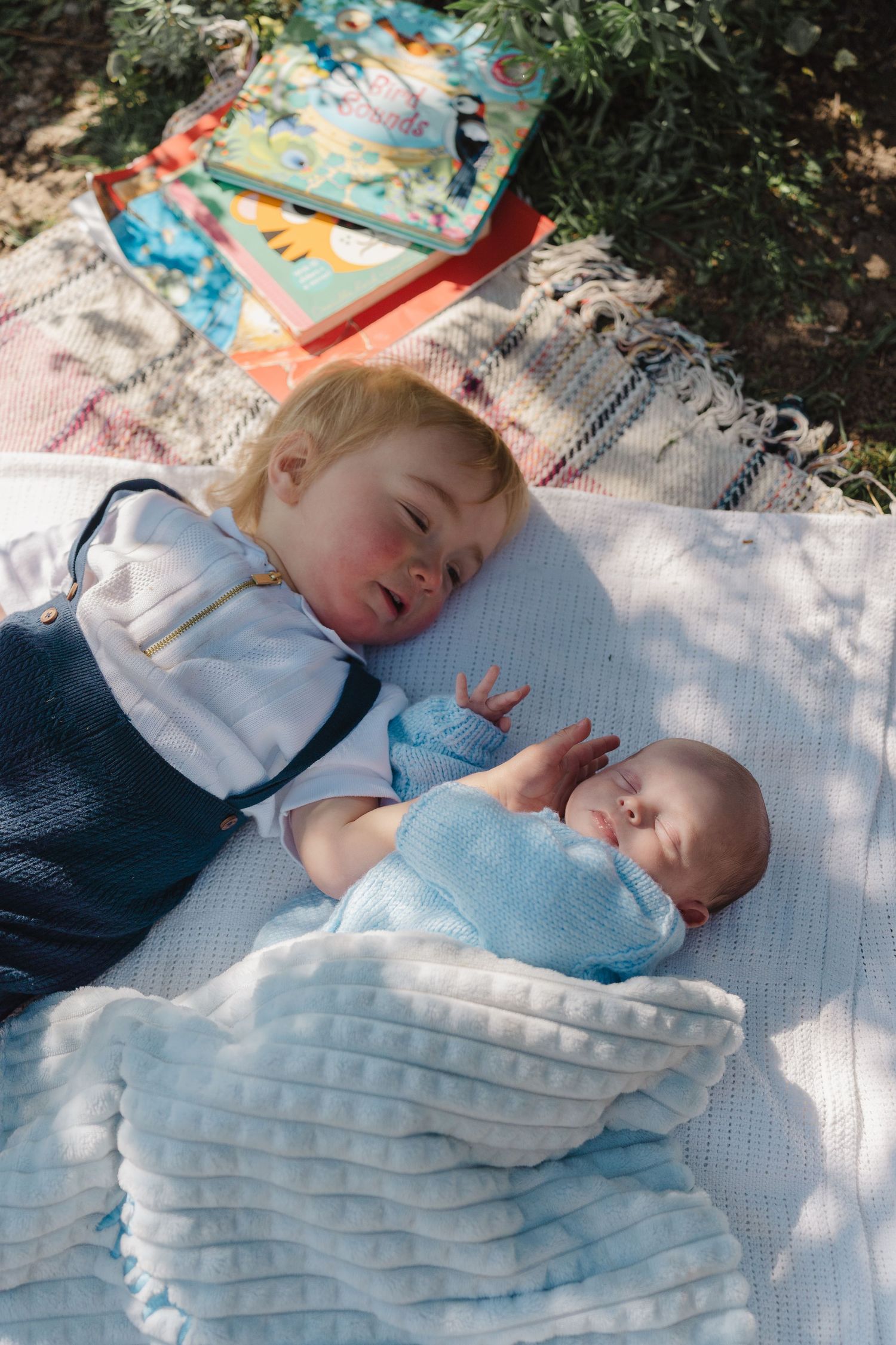 Two children resting on a picnic blanket in the dappled sunlight under trees.