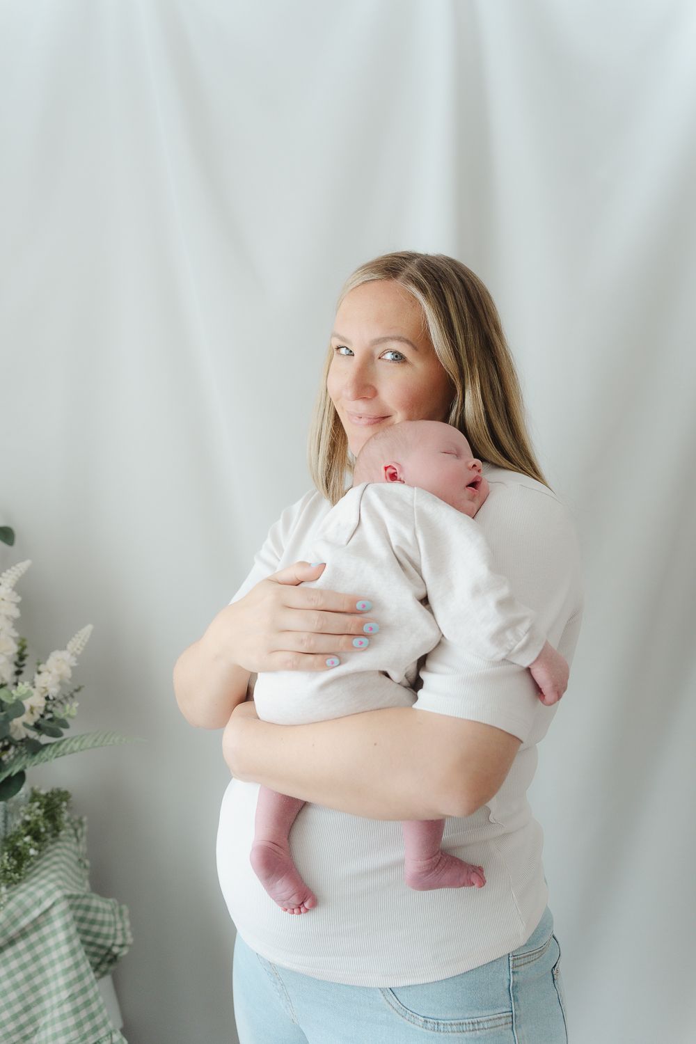 Mother holds baby in white outfit while standing against light background with plants.