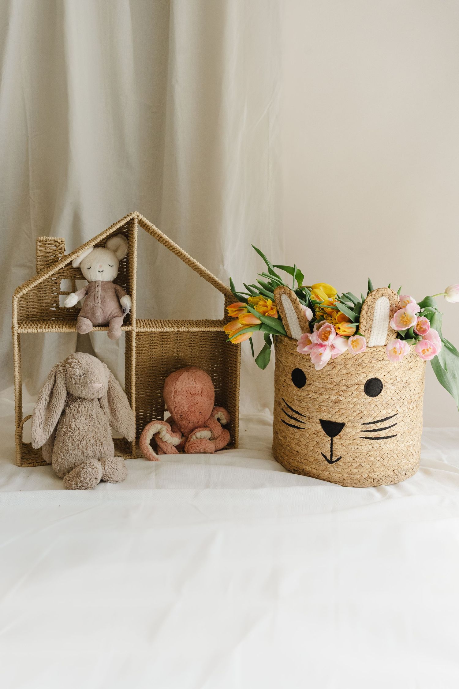 A collection of soft plush toys and decorative baskets arranged in a minimalist white setting with floral accents.
