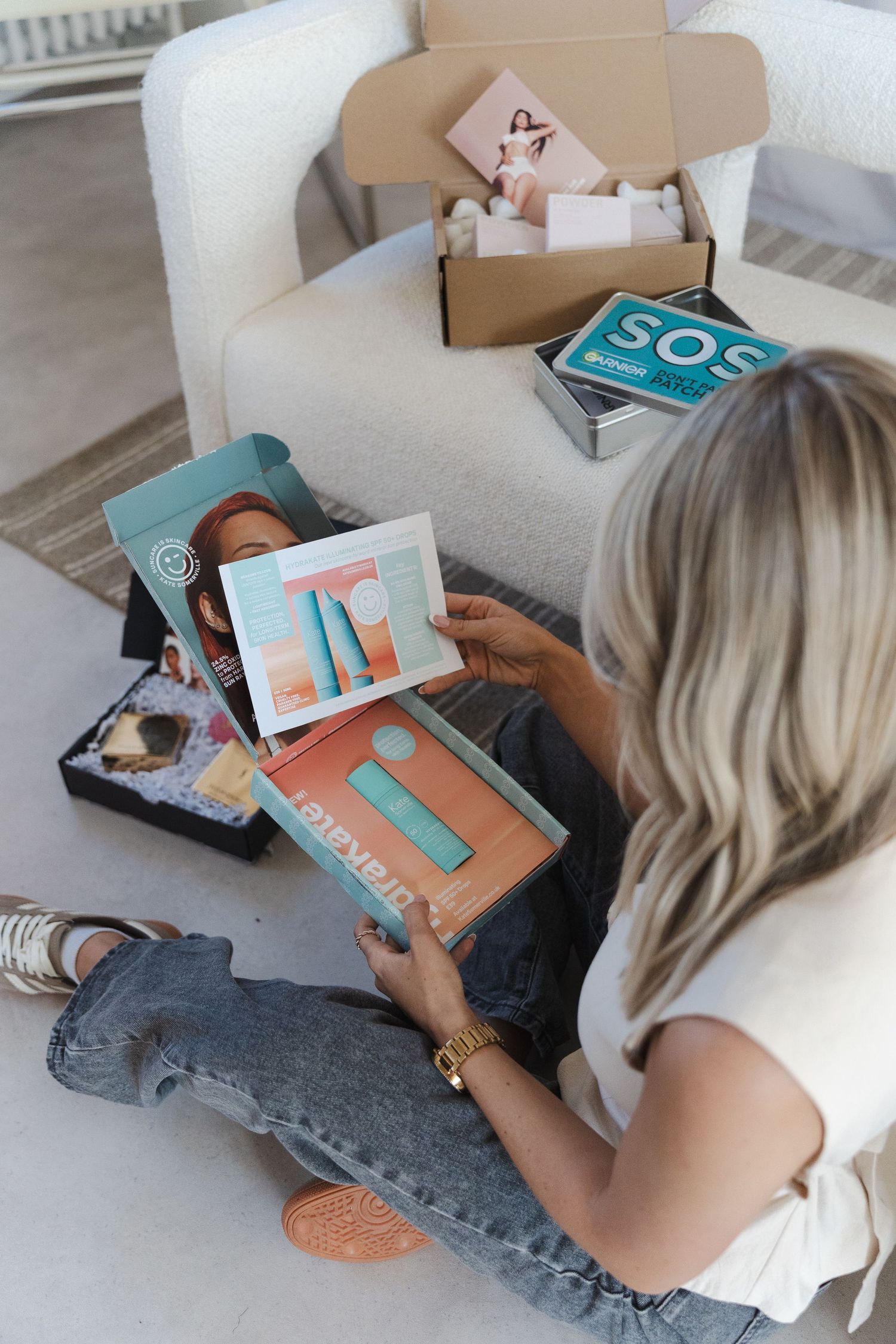 Person lying on white couch opening and examining subscription box contents in casual home setting.
