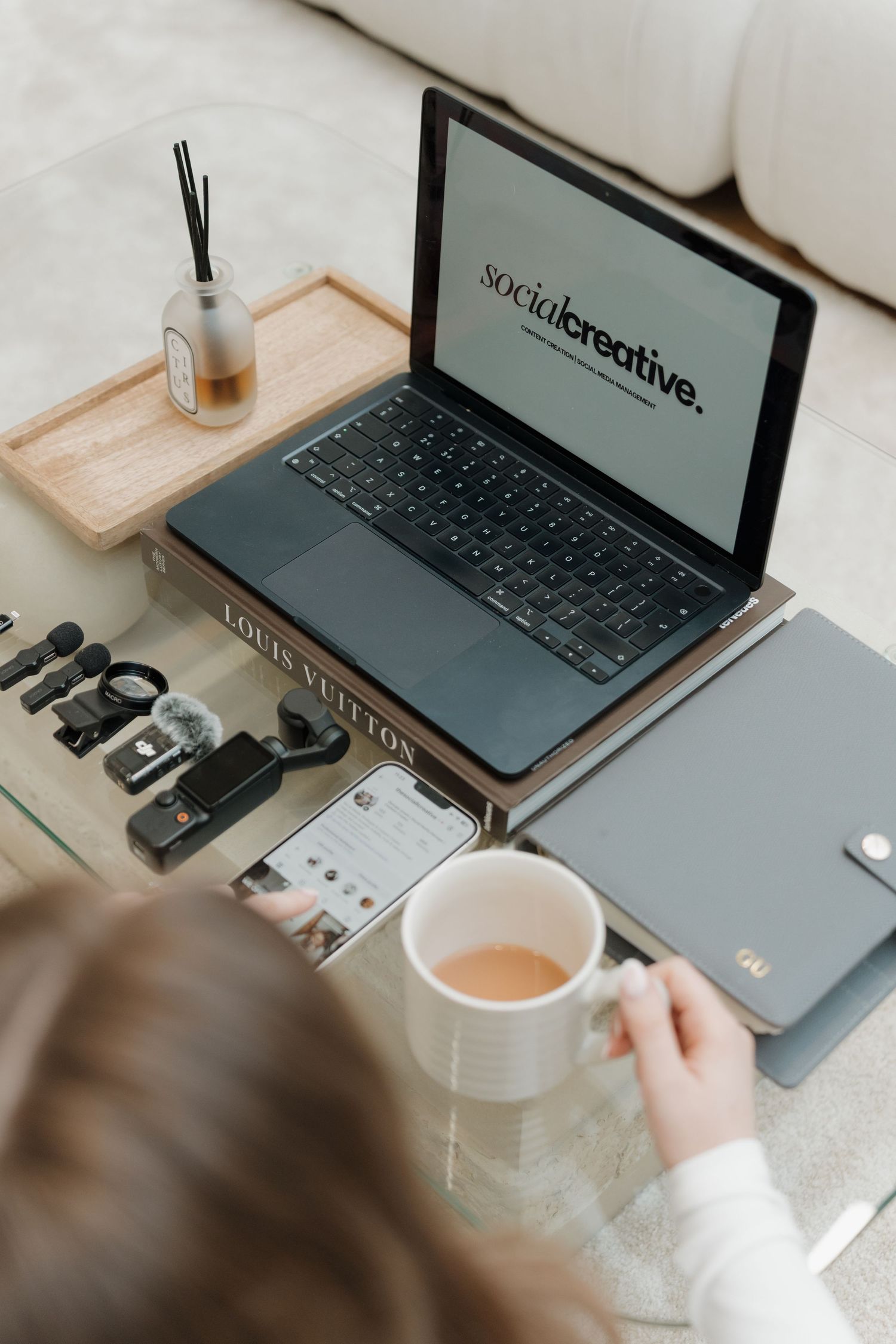Person holding a coffee mug while working on a laptop displaying Social Creative website at a stylish home office desk. Content Brand Day.