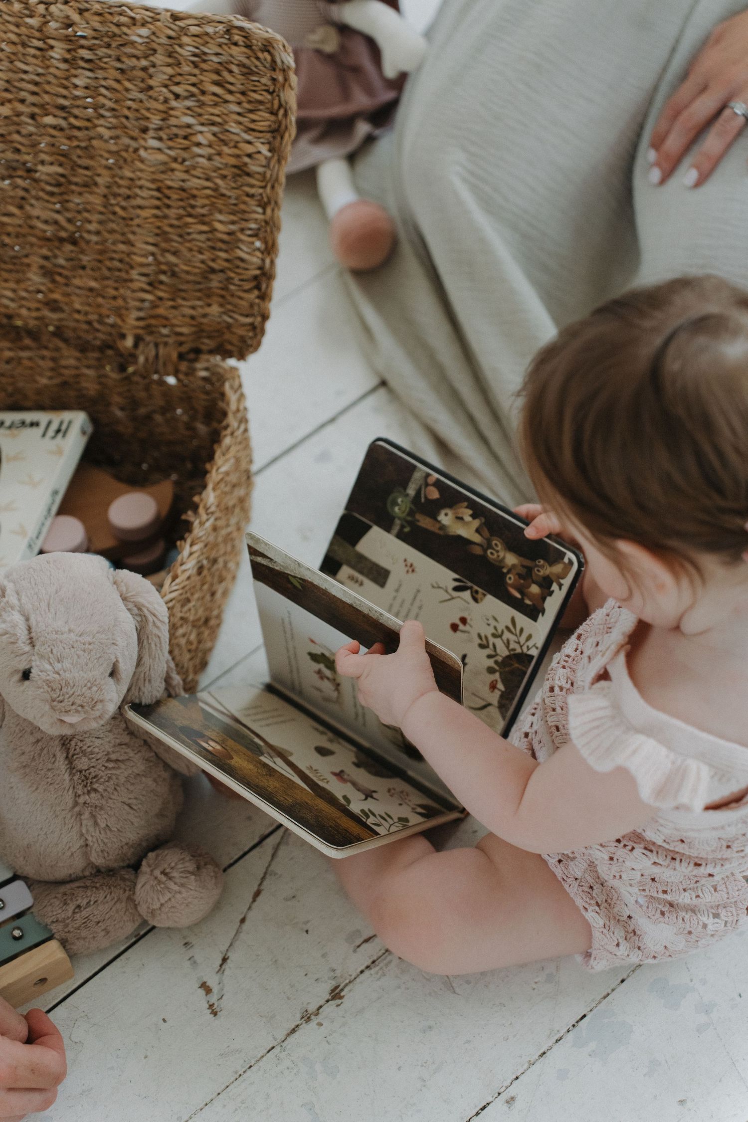 Toddler girl in floral dress sits on white floor reading a book beside a wicker basket and stuffed bunny toy.