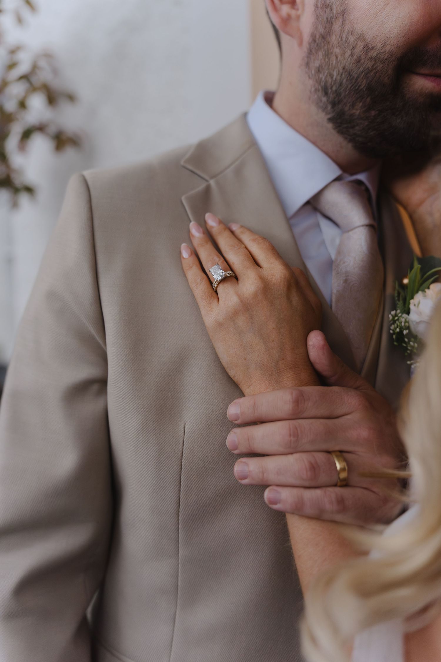 Close-up of bride and groom hands showing diamond engagement ring and gold wedding band on beige suit.