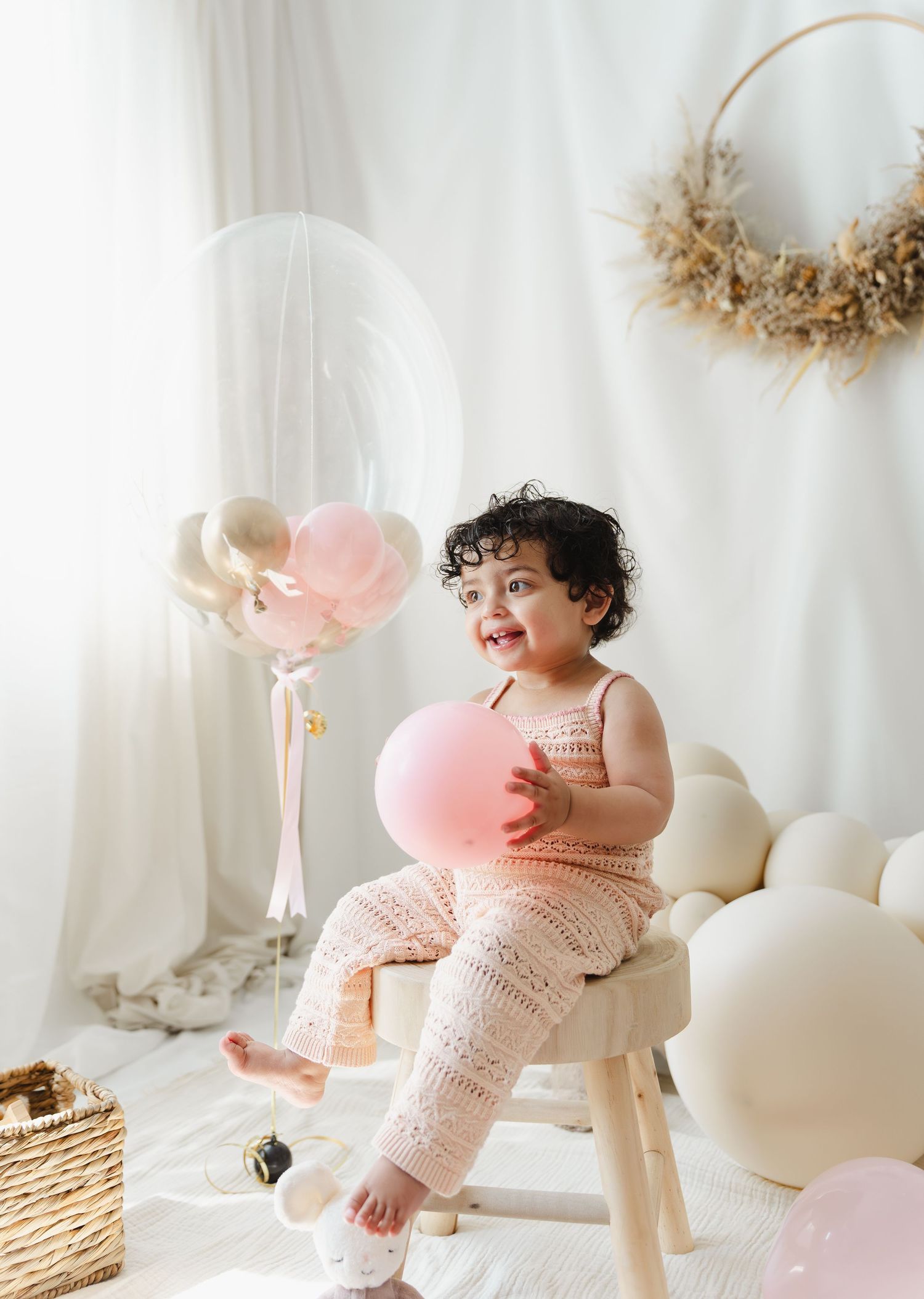 Sweet baby in pink knit dress laughs while playing with pink balloons in a bright white studio setting.