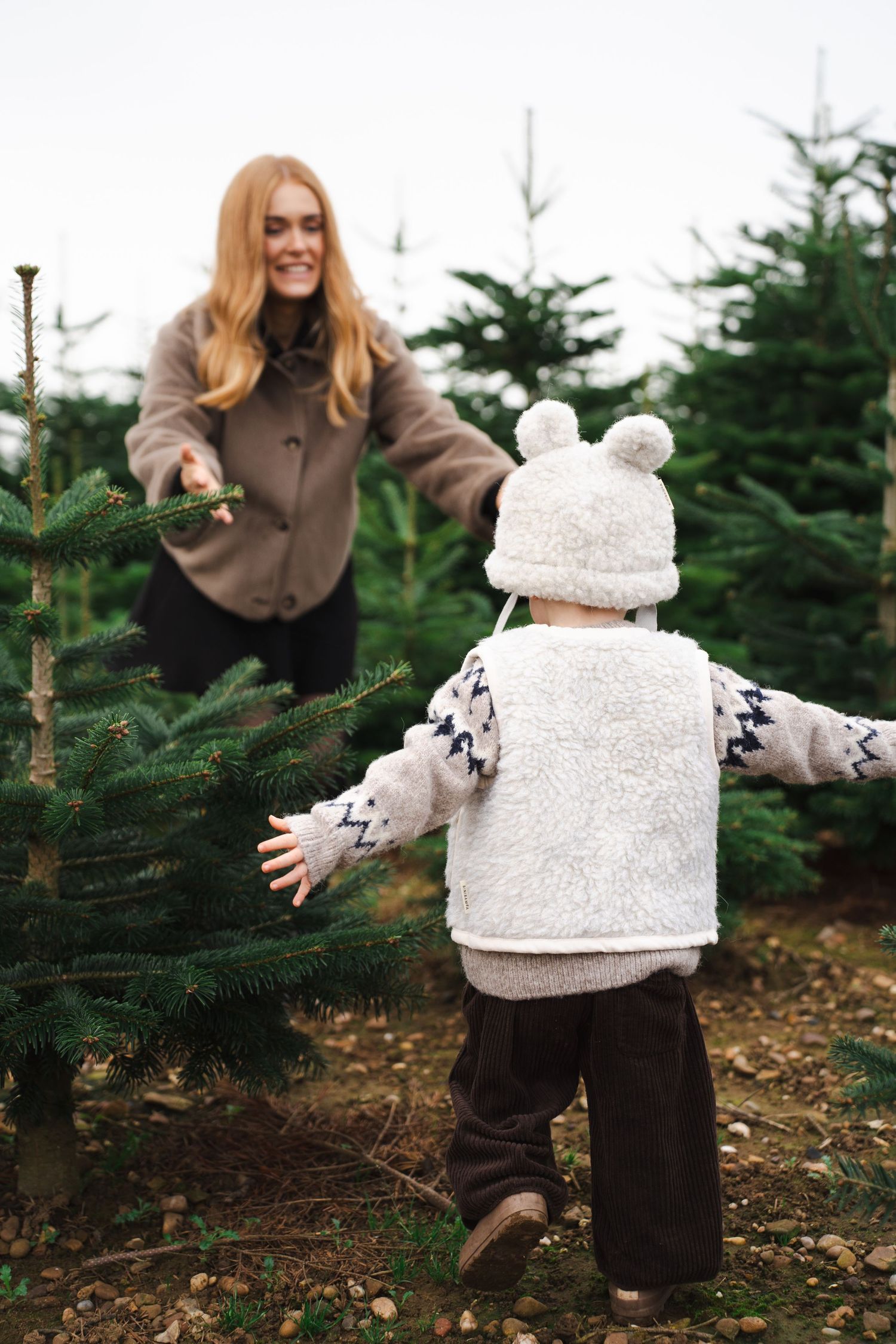 A parent and child in winter coats explore together among evergreen trees at a Christmas tree farm.