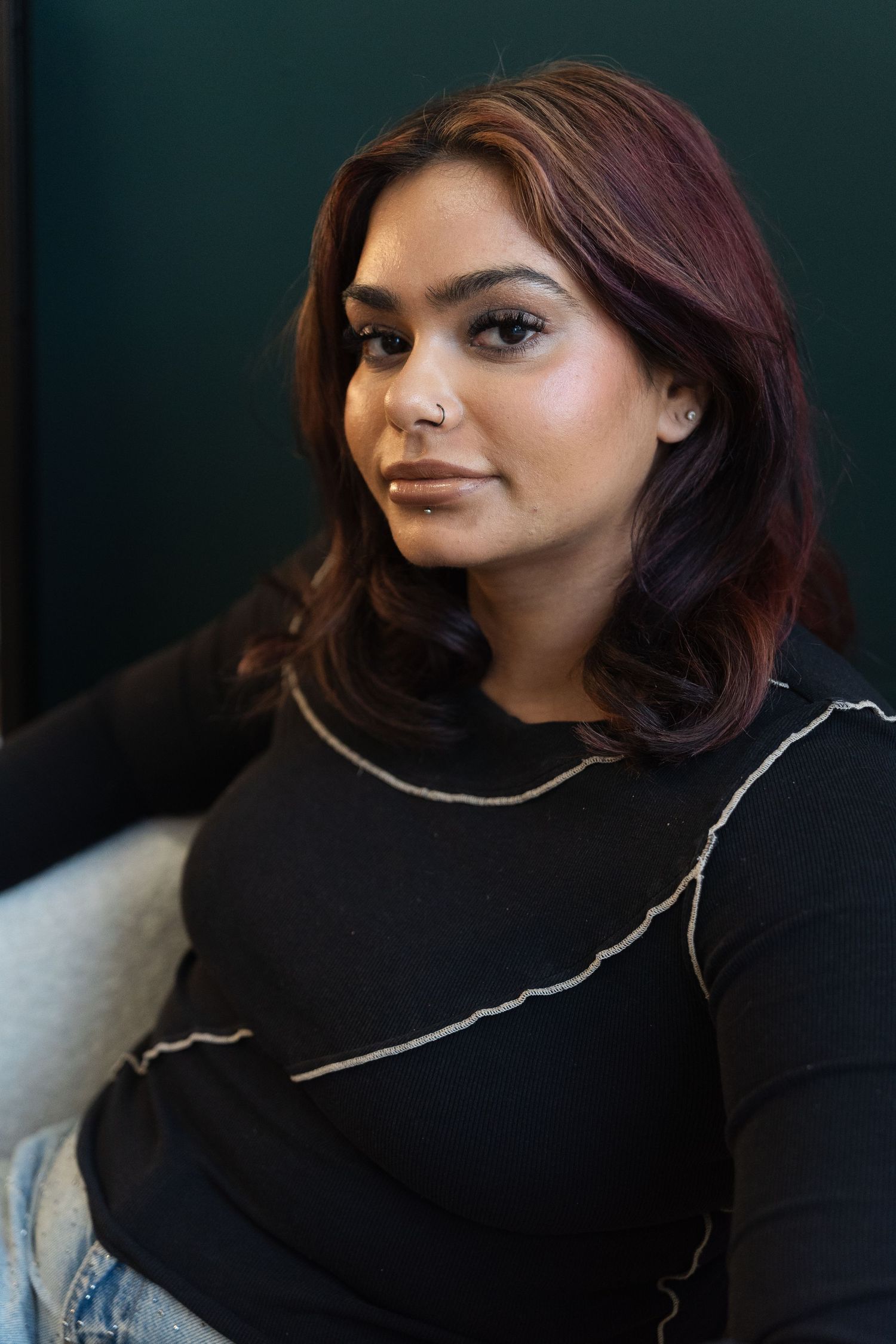 Portrait of someone with burgundy-tinted hair wearing a black sweater with white trim against a dark green background.