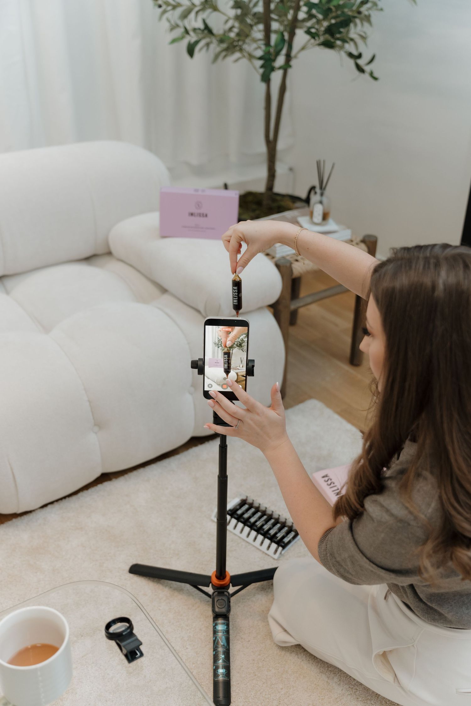 Woman adjusting smartphone on tripod to record beauty content in a cozy white living room setup.
