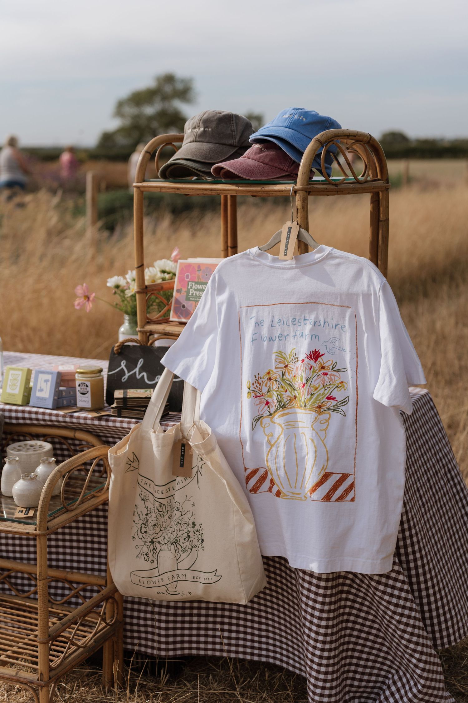 White floral t-shirt and tote bag displayed on rattan stand at outdoor market stall with caps and gingham tablecloth.