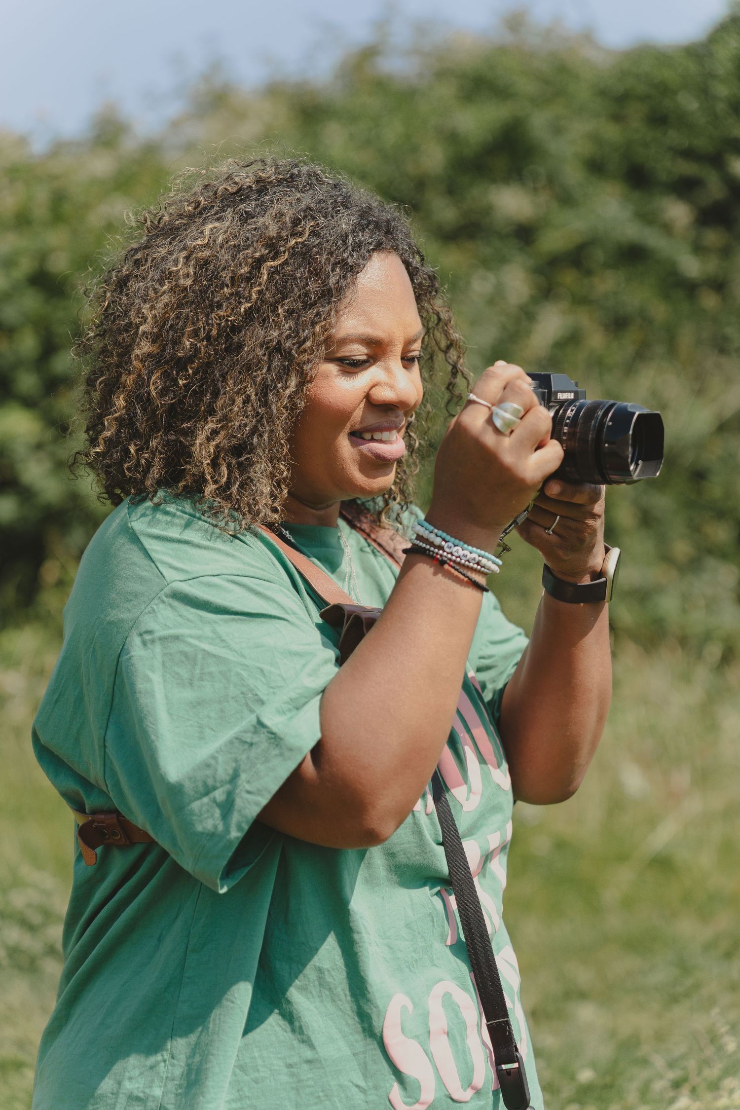 Photographer with curly hair wearing a mint green shirt smiles while taking pictures outdoors on a sunny day.