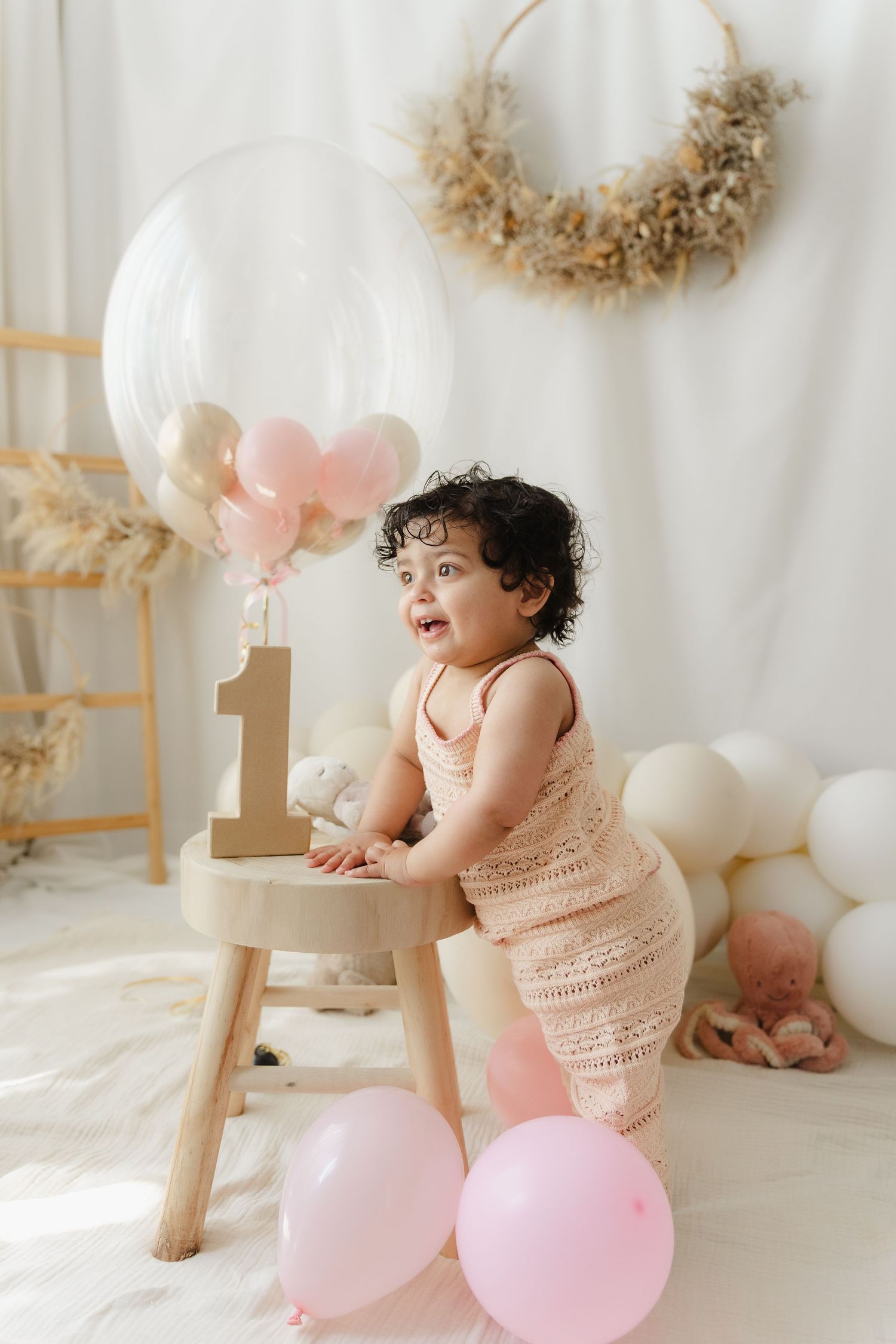 Baby in pink dress sits on wooden chair surrounded by pink balloons and dried floral decorations.