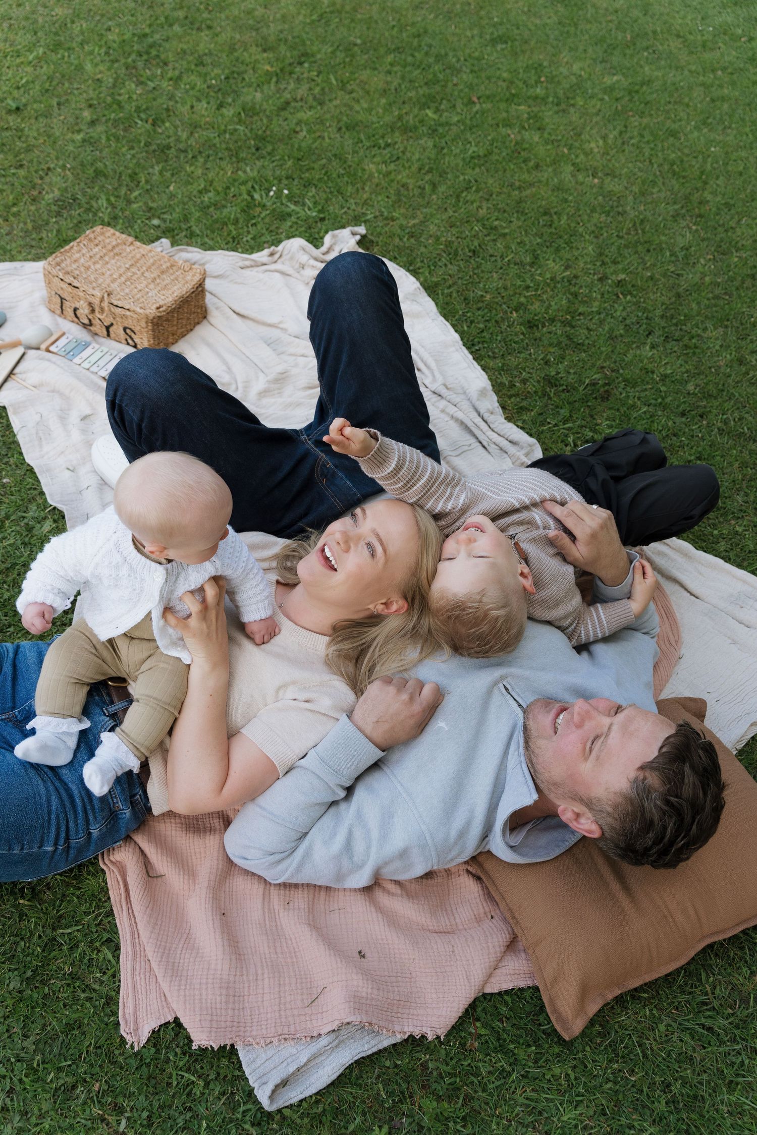 Happy family with baby and toddler relaxing on a blanket in green grass during a sunny outdoor picnic.