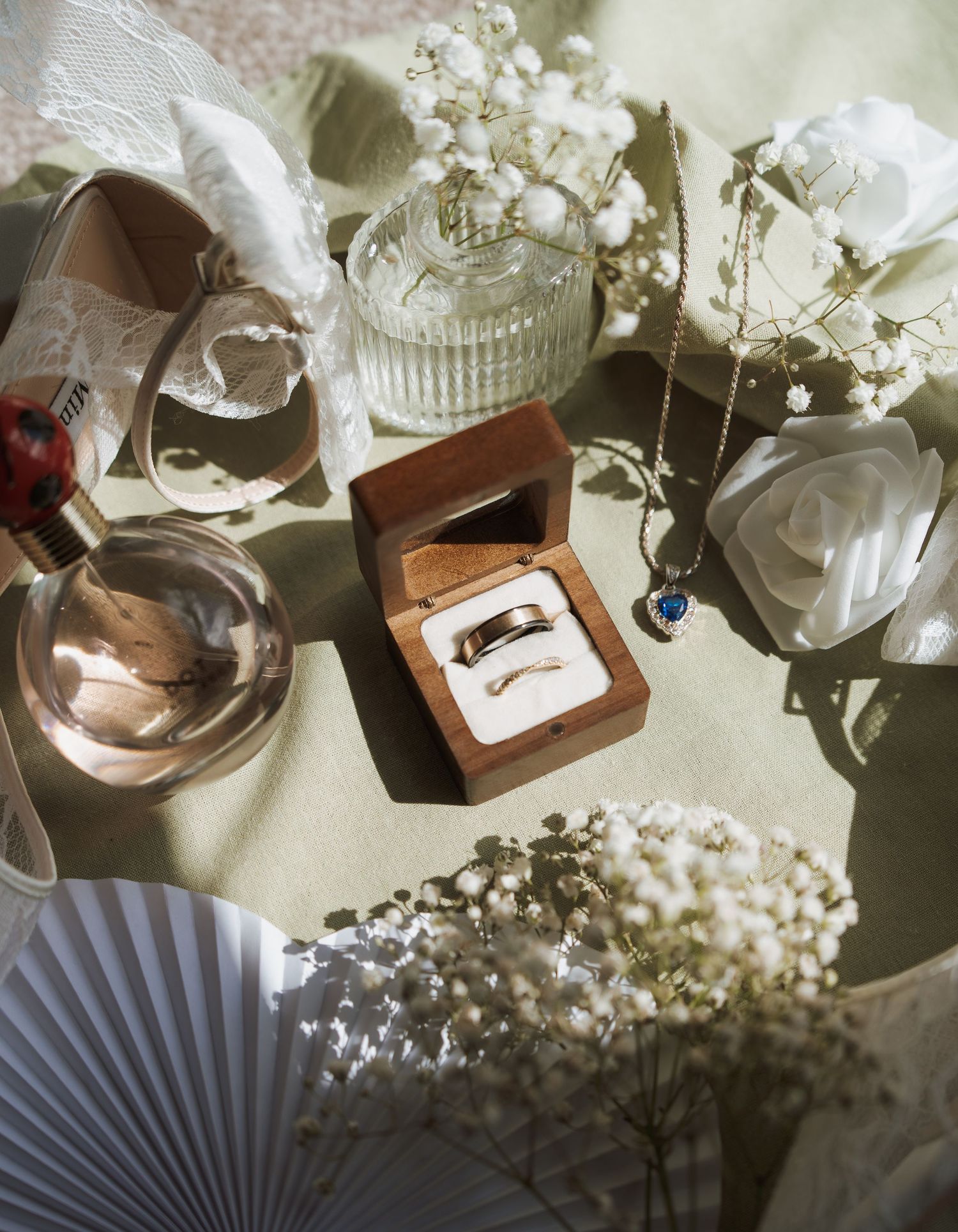 Elegant wedding flat lay with gold rings in wooden box, white flowers, perfume, and blue heart necklace on green surface.