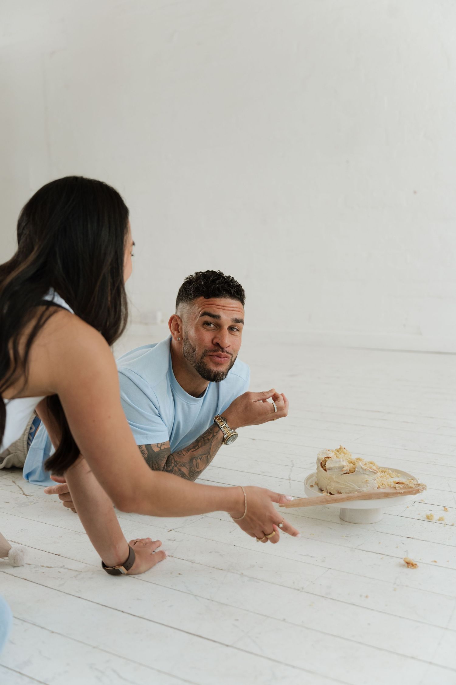 A man and woman sit on a white floor sharing a cake, with the man looking up while eating a piece.