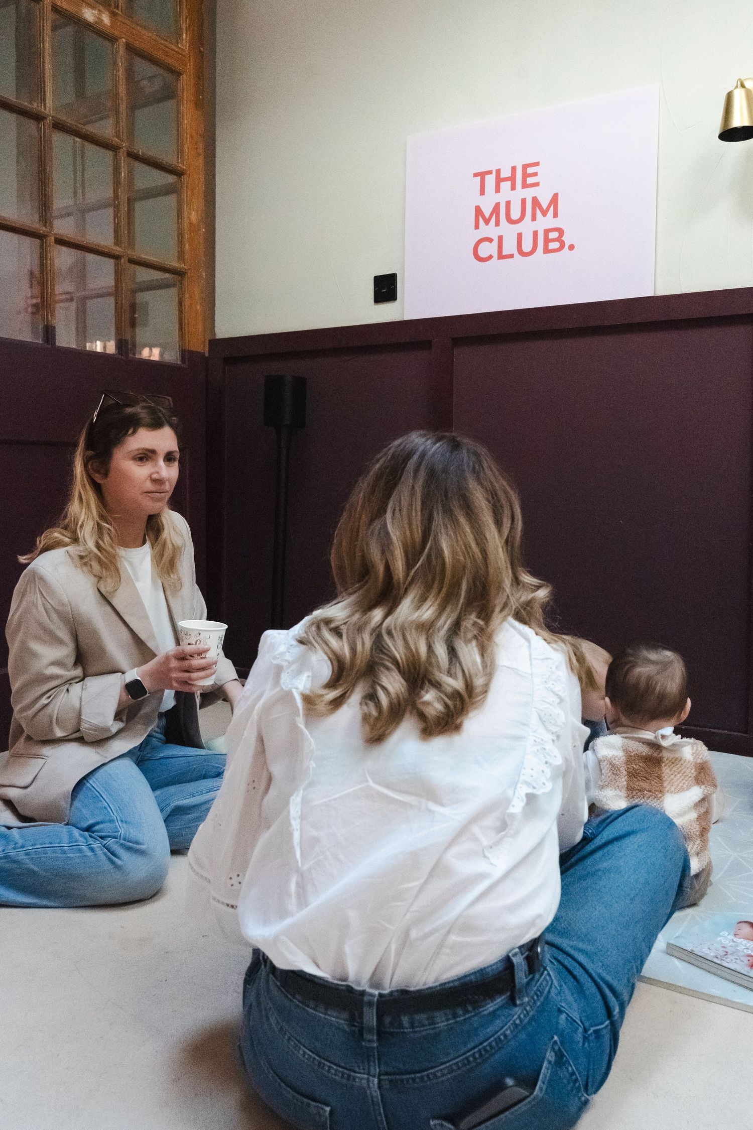 Two people sit on floor against white wall with pink text signage in modern minimalist space.