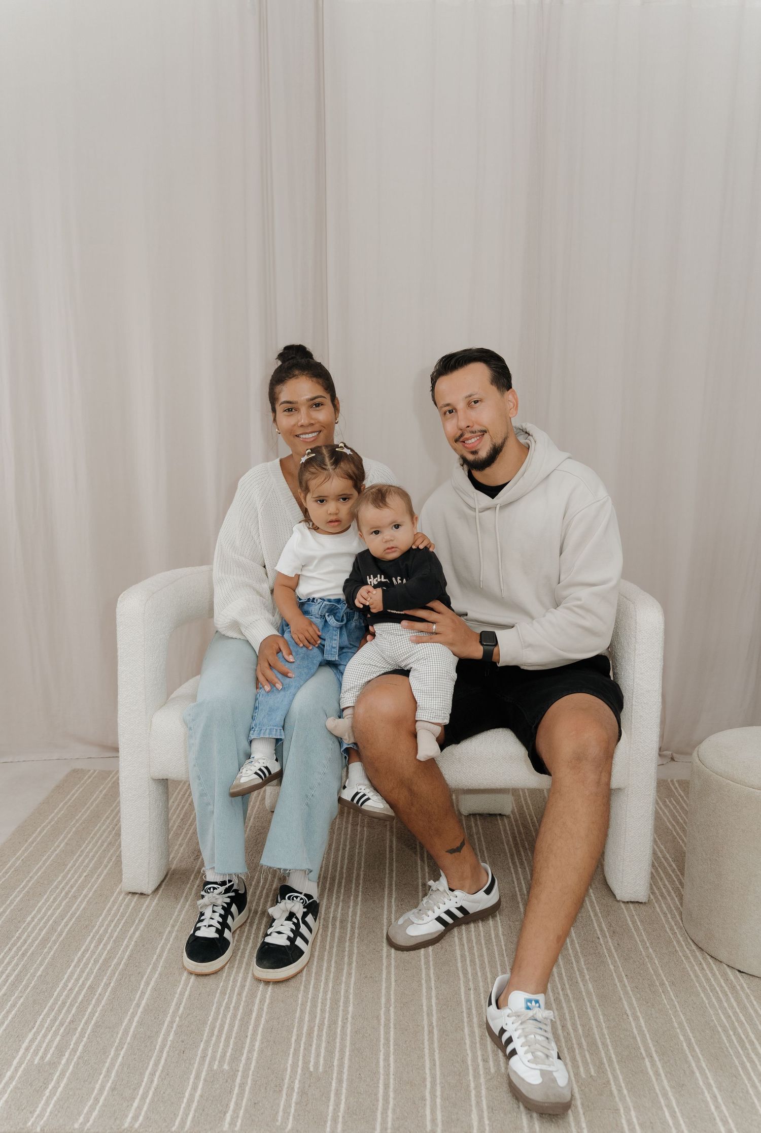 A family wearing white and casual attire sits together on a white chair against a light curtain backdrop.