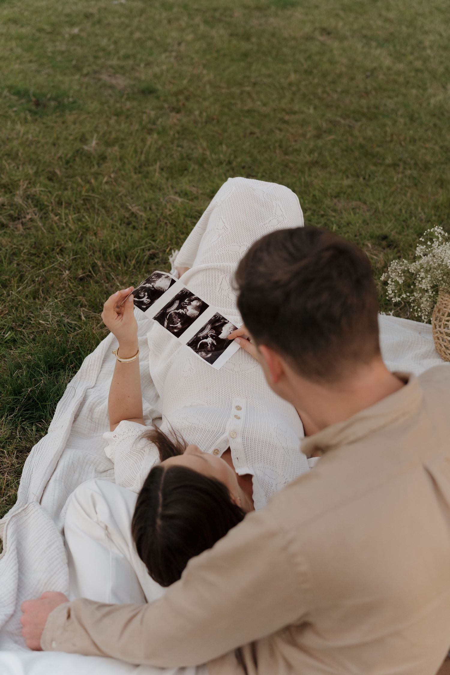 A couple sitting on a white blanket outdoors looking at ultrasound photos together on grass.