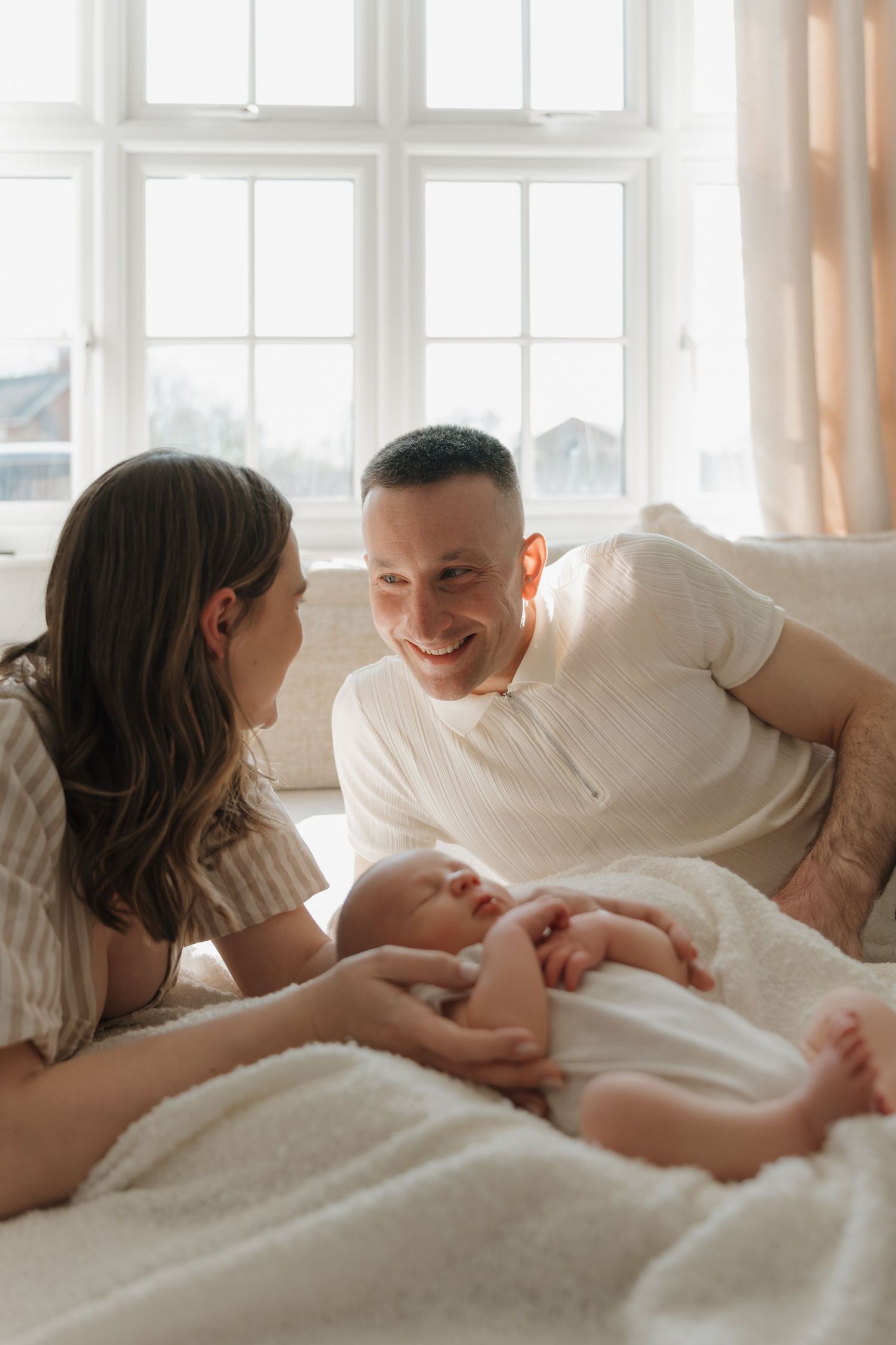 Happy parents smiling at their newborn baby lying on a white blanket in a bright, sunlit bedroom. Home Newborn Photography Derbyshire.