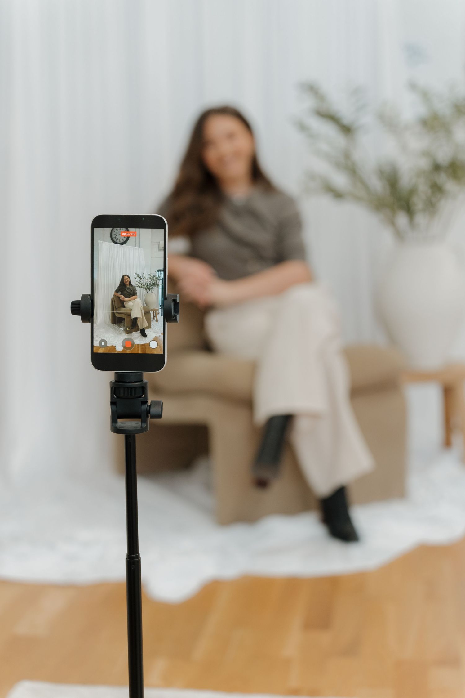 Woman content creator recording herself on a smartphone mounted on a tripod in a modern minimalist studio setting.