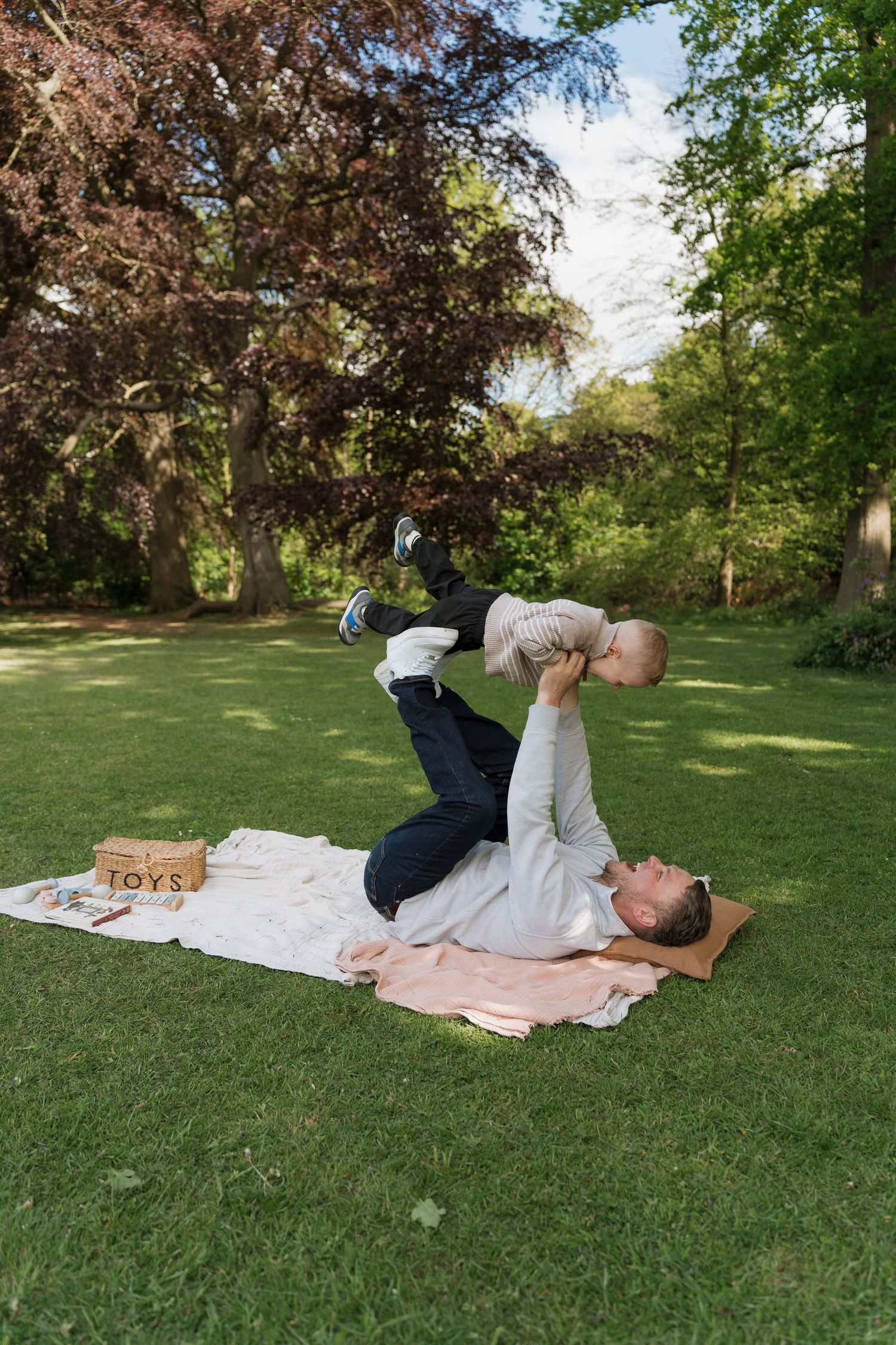 Parent lying on a picnic blanket in a lush park, playfully lifting a baby overhead on a sunny day.