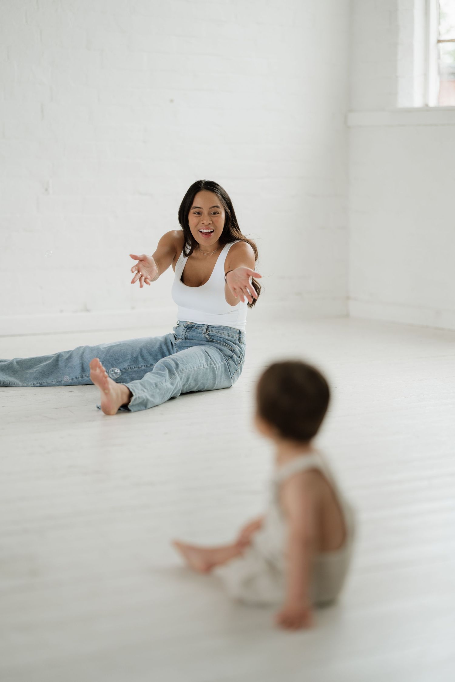 A laughing mother sits on a white floor, reaching out to her baby sitting nearby in a bright white room.