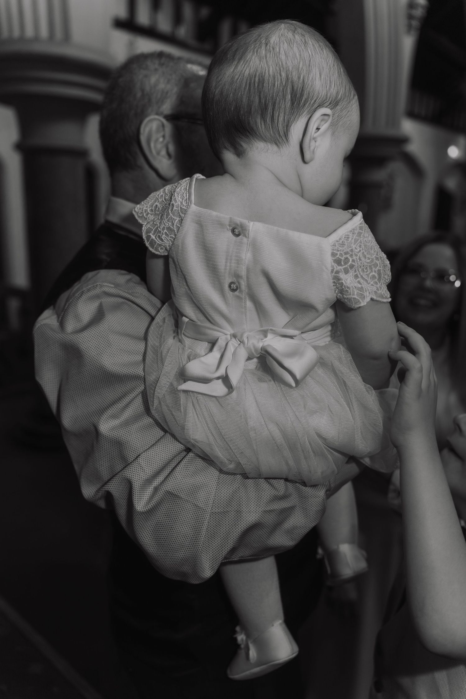 Black and white photo of a baby wearing a formal white dress with lace details and a bow at the back.