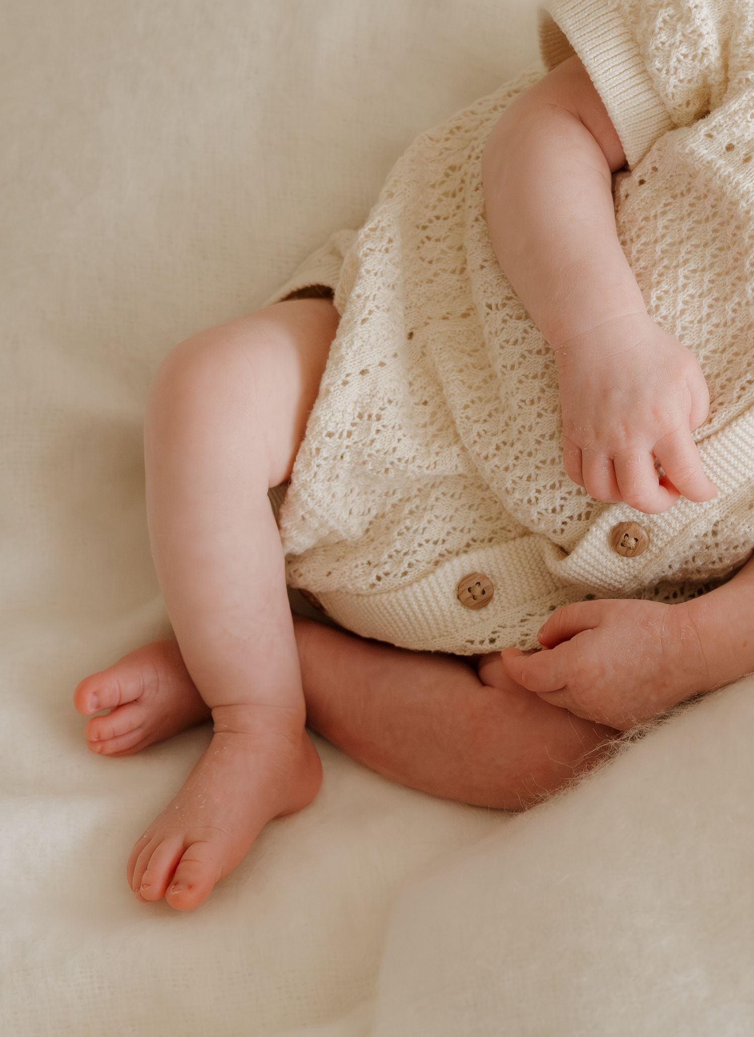 Close-up of tiny baby feet and legs wearing a cozy cream-colored knit romper with buttons on white bedding.
