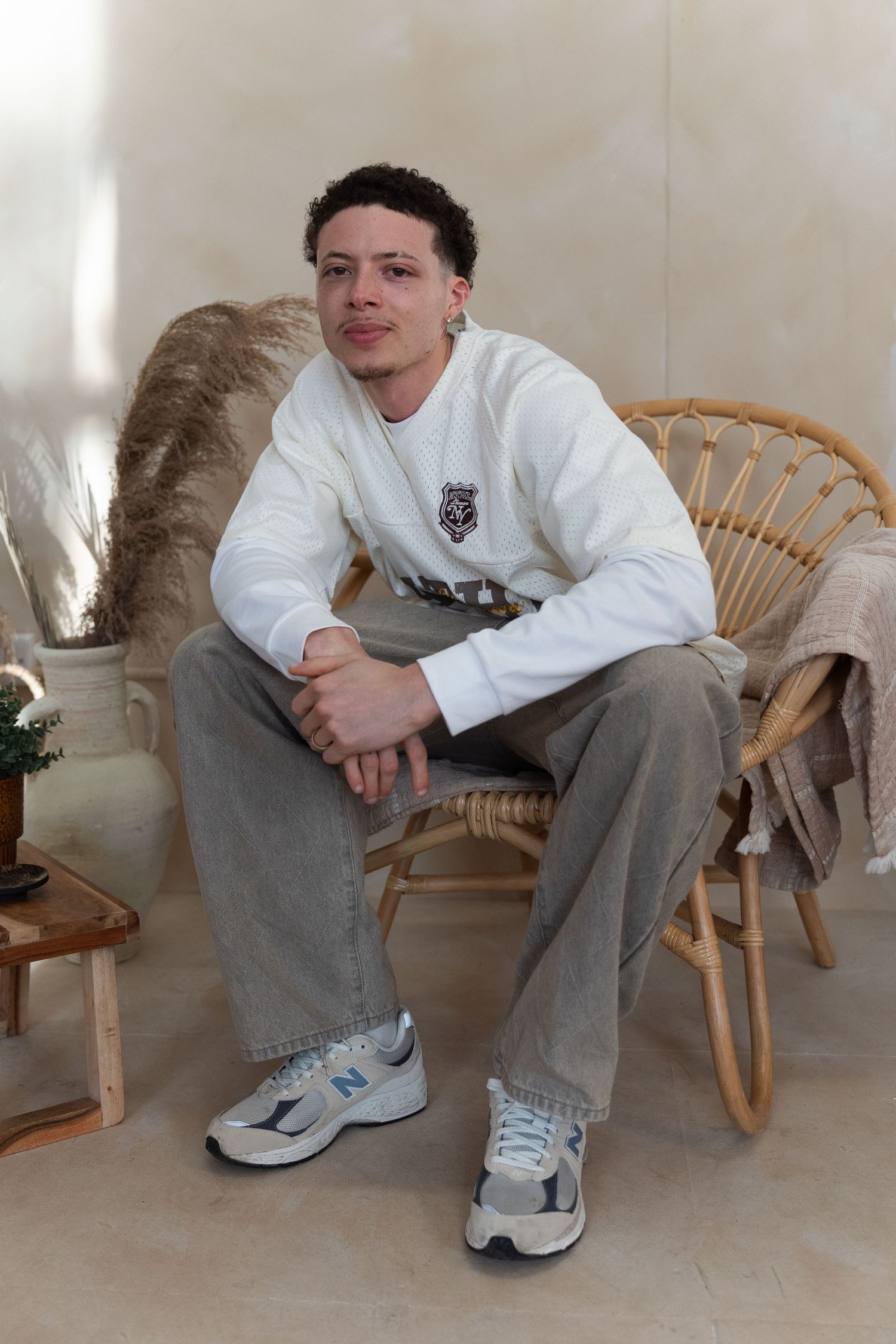 A person in a white sweatshirt and gray pants sits casually on a rattan chair with pampas grass decor in the background.