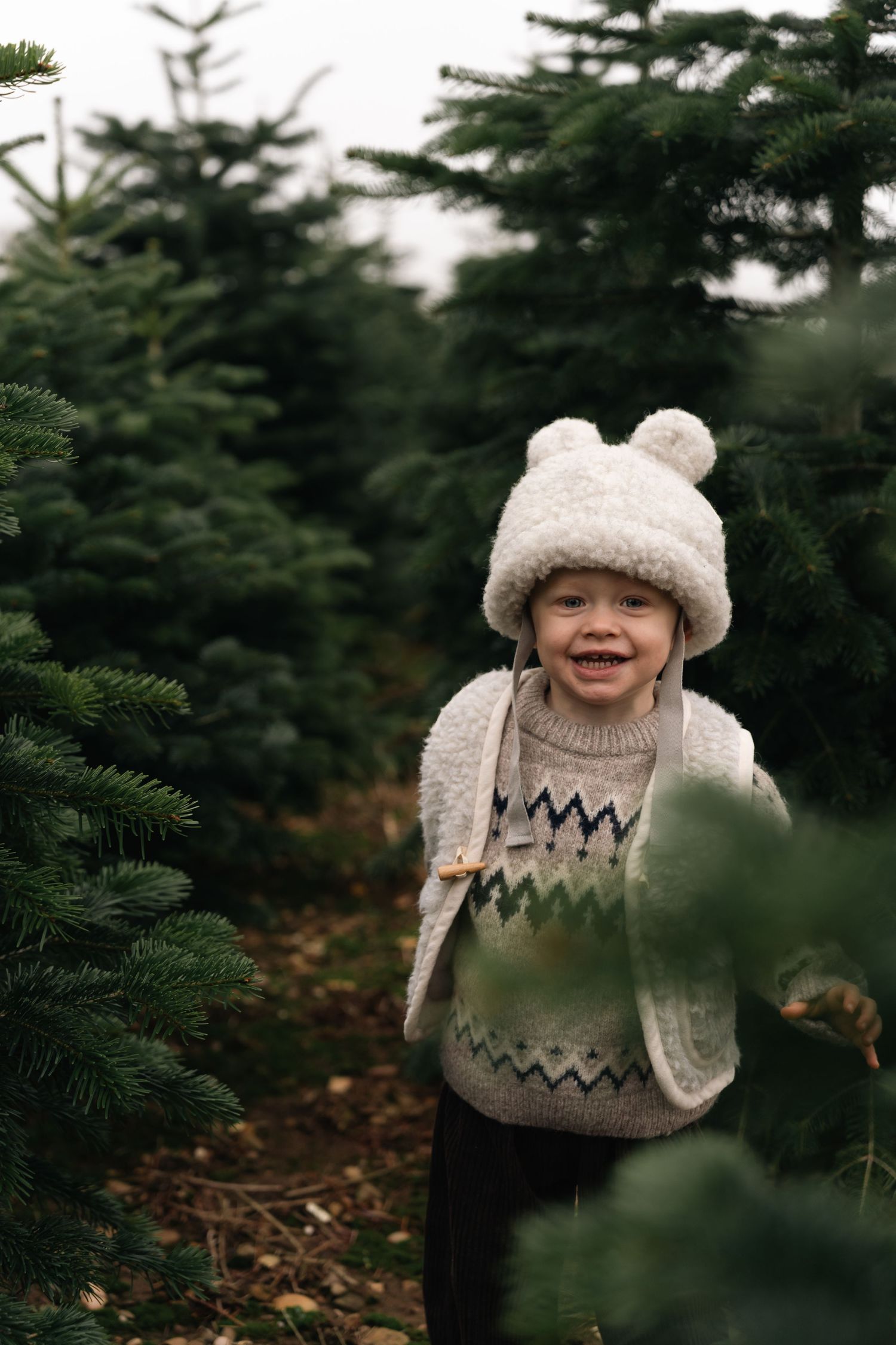 Smiling toddler in white bear hat and knit sweater exploring a Christmas tree farm.