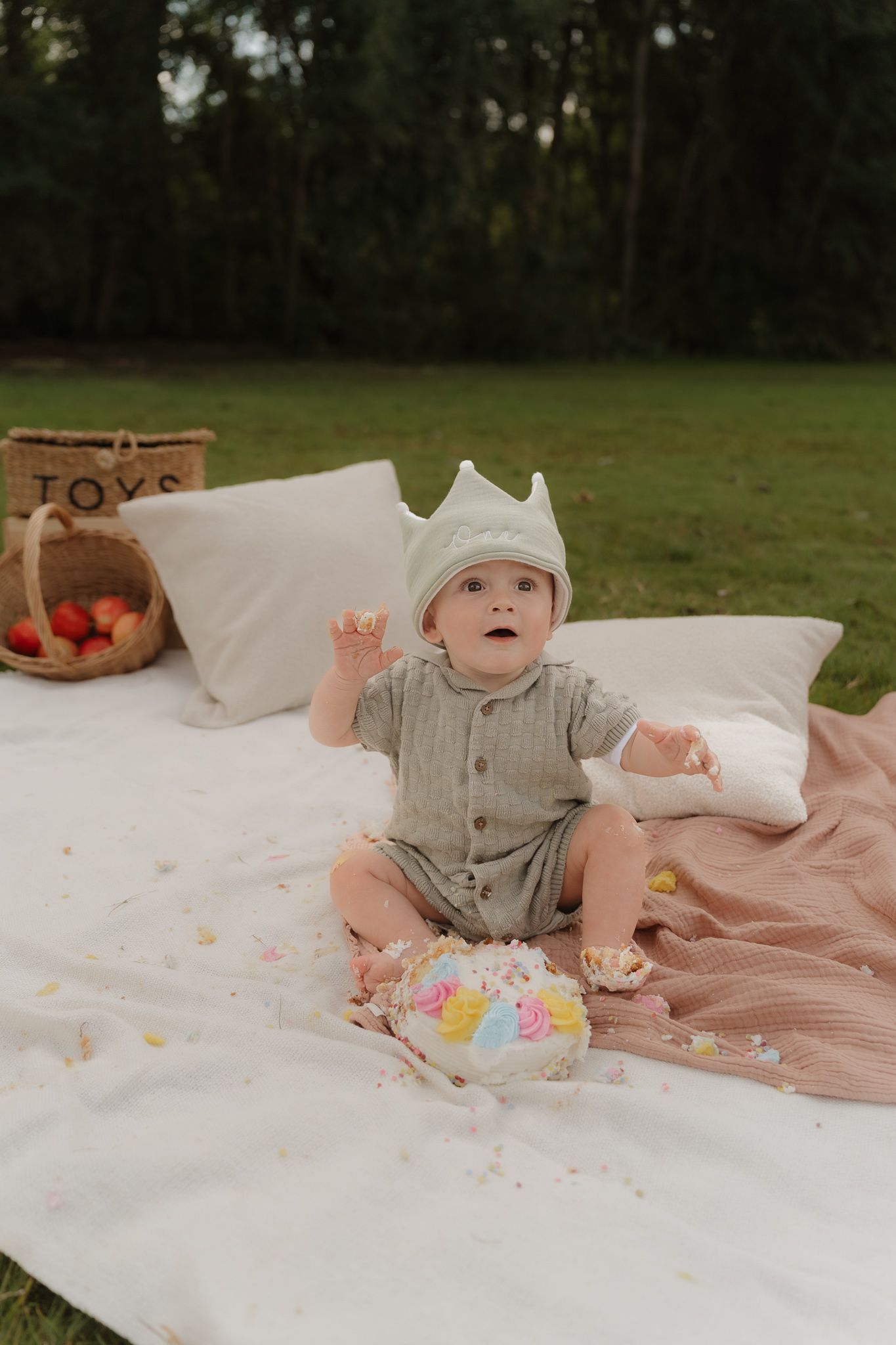 Baby's first birthday celebration setup outdoors with cake and decorations on white blanket.
