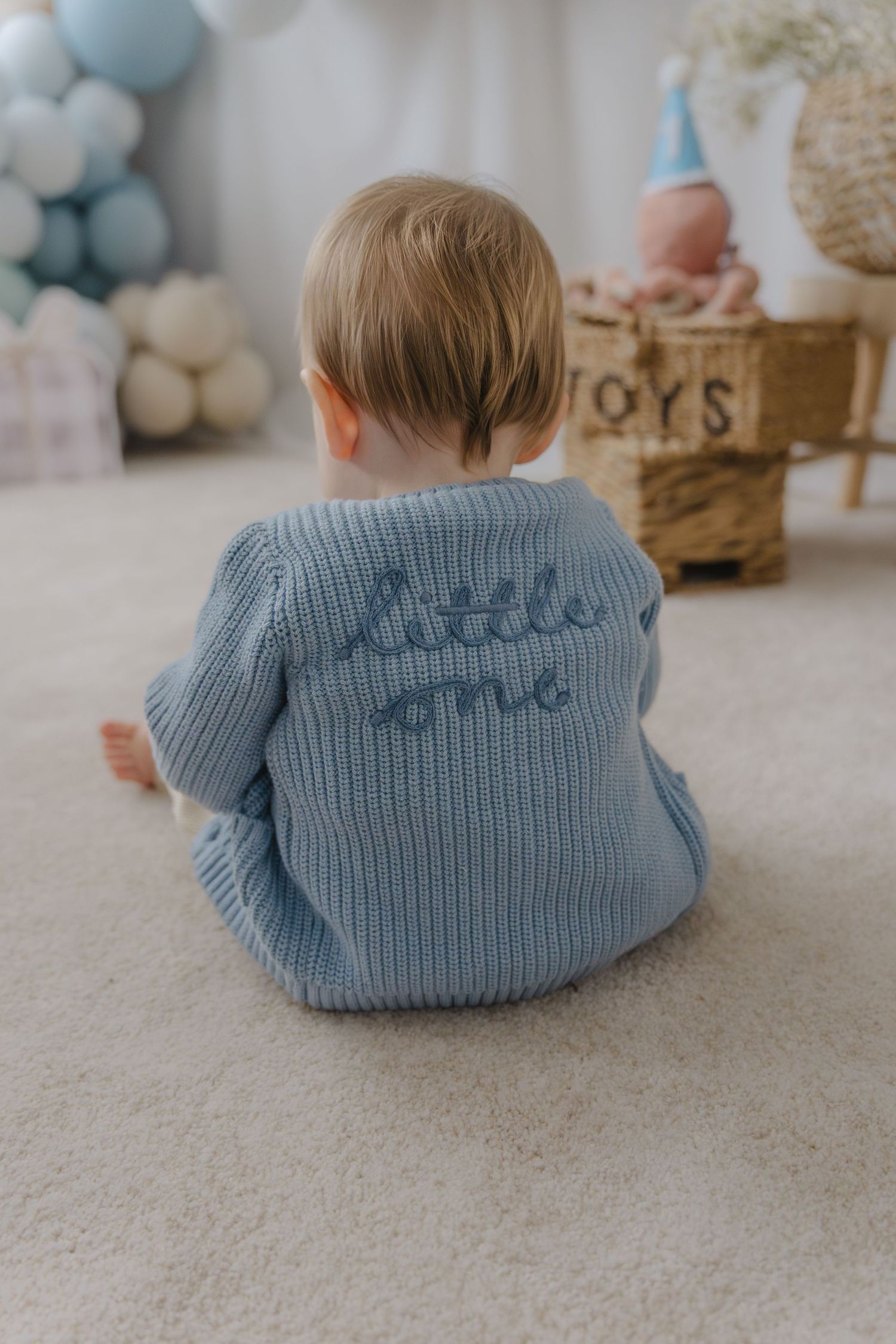 Baby wearing blue knit sweater with text detail sitting against balloon decorated background.